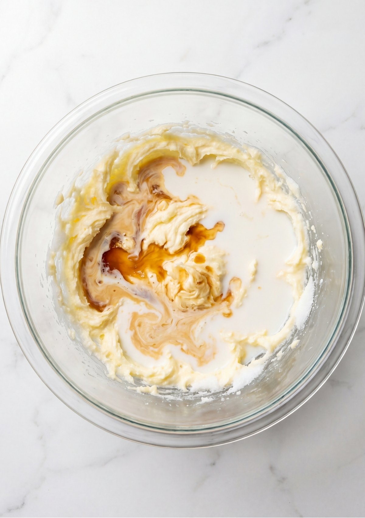 Vanilla extract and milk being mixed into creamed butter and sugar in a clear glass bowl