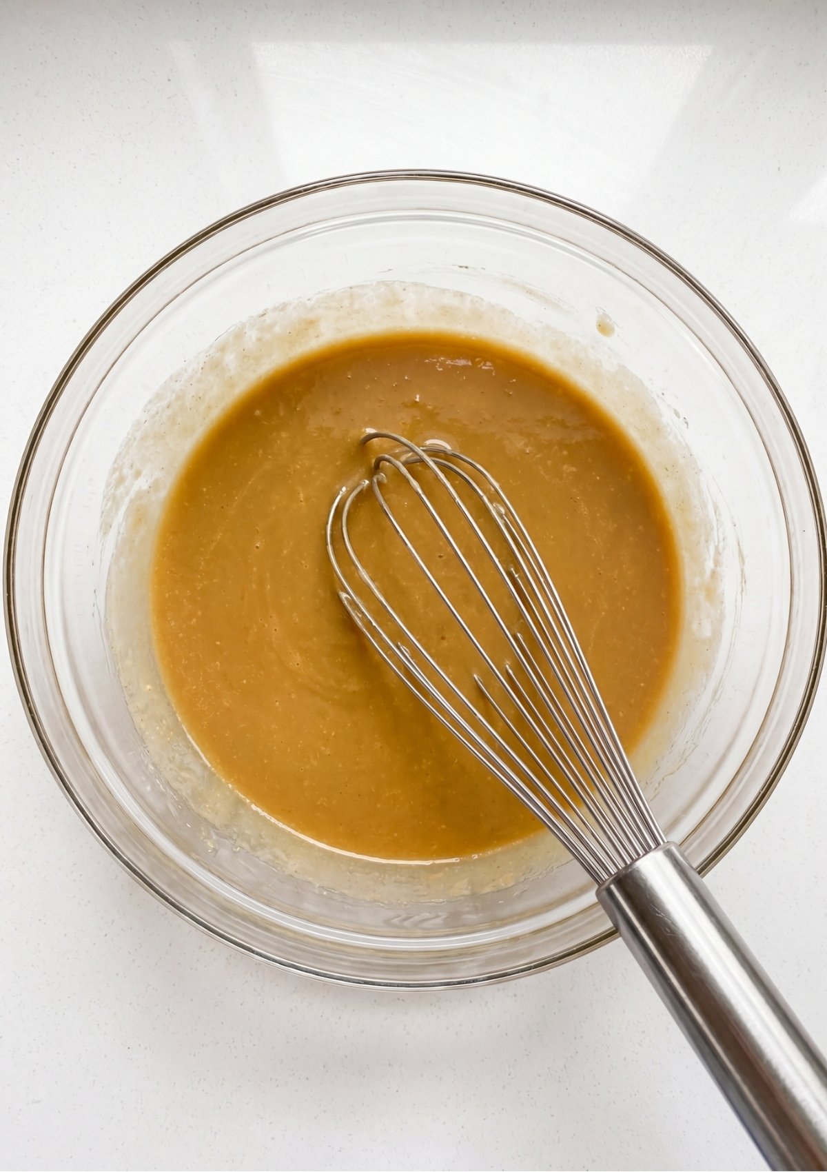 A metal whisk resting in a smooth glossy mixture of melted butter and sugars inside a glass bowl