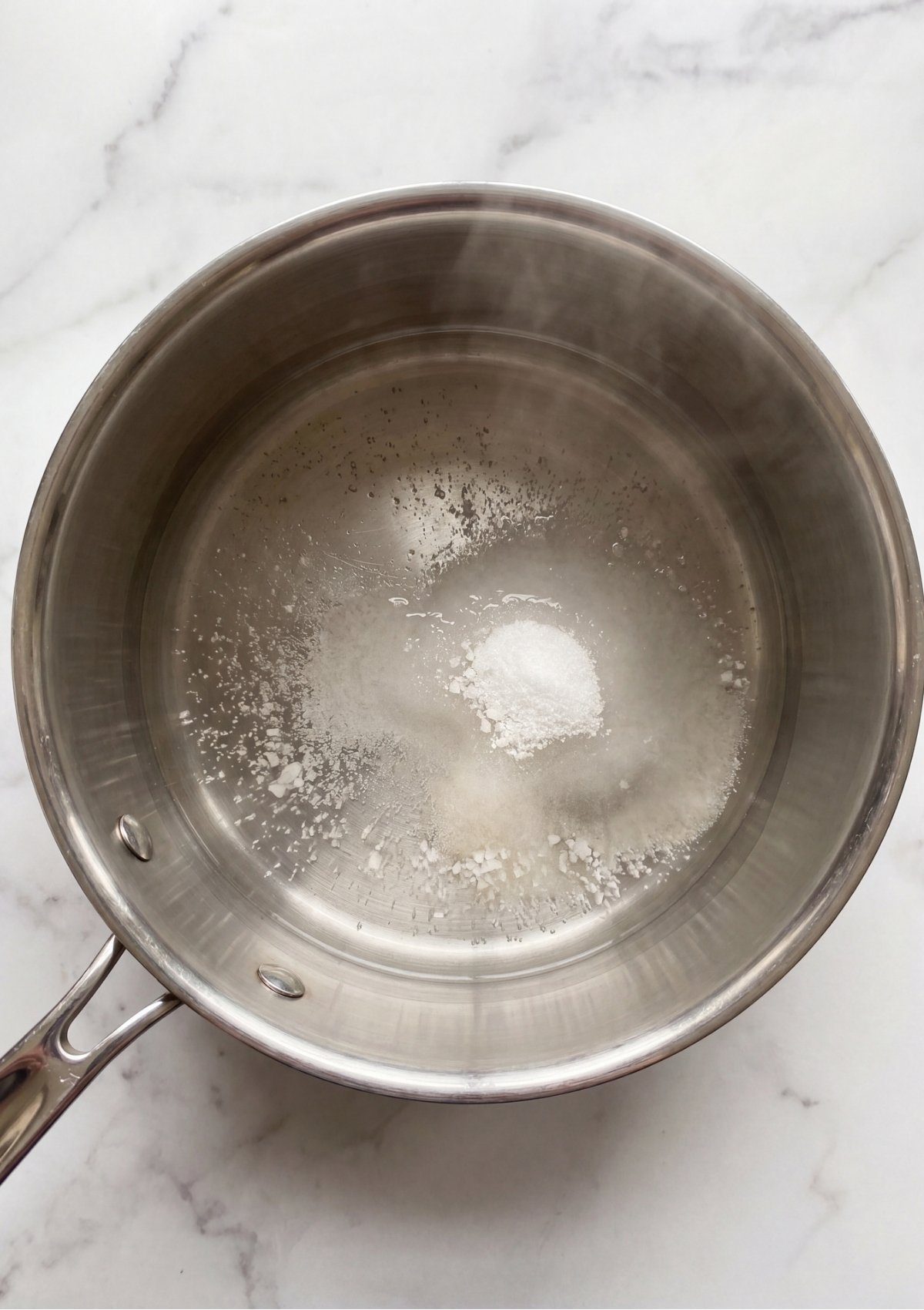 Cane sugar and sea salt dissolving in a simmering water and vinegar brine inside a stainless steel saucepan on the stovetop.