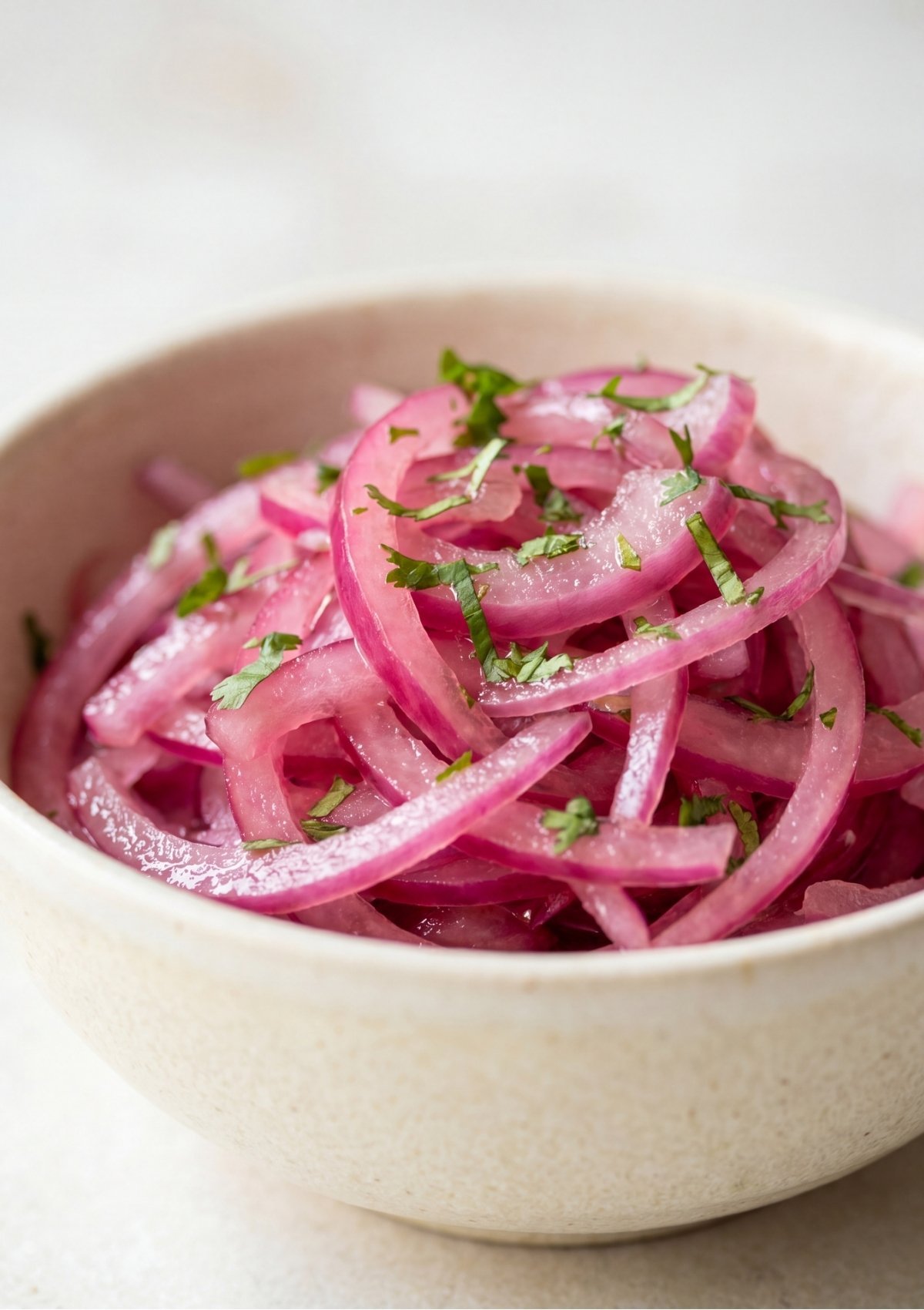 A close-up shot of a beige ceramic bowl filled with bright pink pickled red onion slices mixed with finely chopped fresh cilantro.