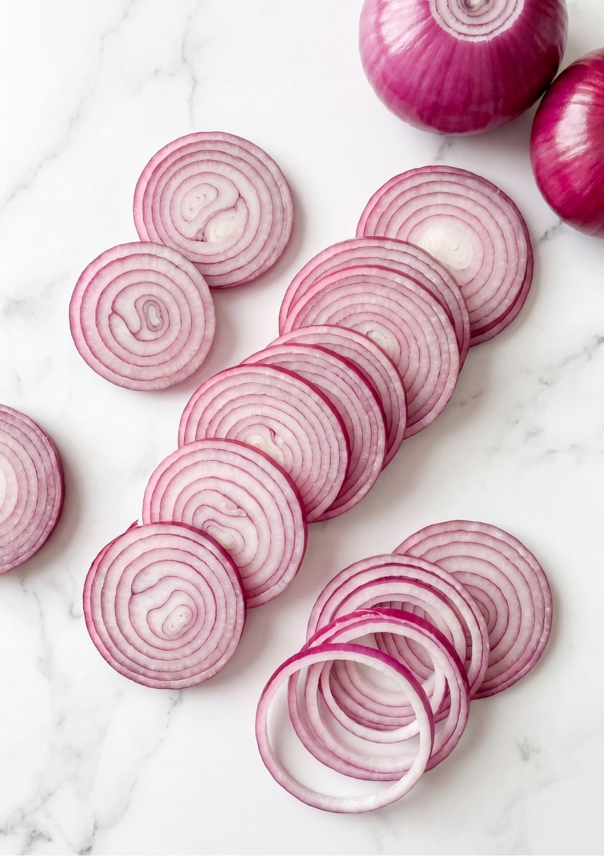 Evenly sliced red onion rings arranged in rows alongside two whole red onions on a white marble tabletop