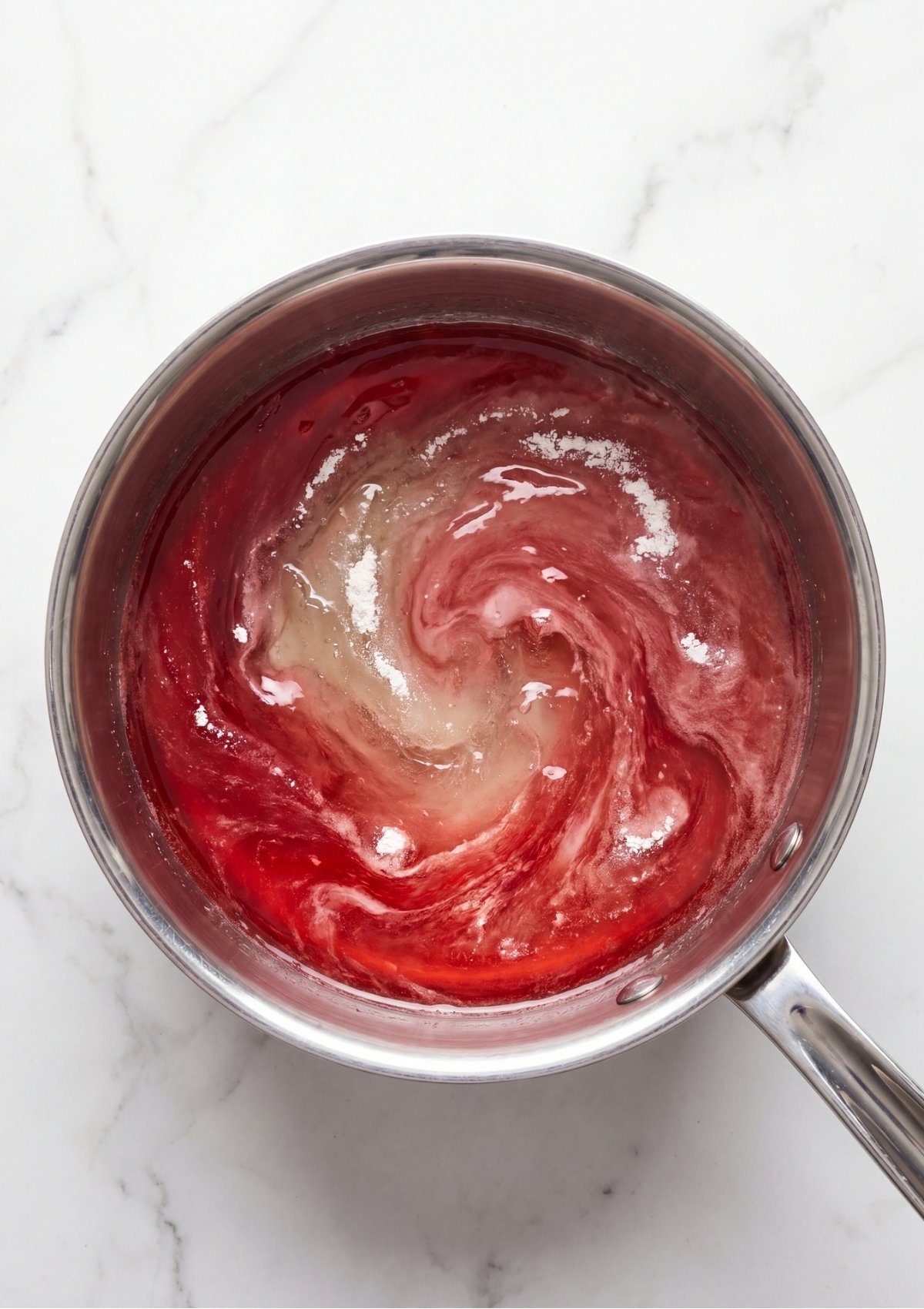 Red gelatin powder and bloomed gelatin mixture being stirred into hot liquid inside a stainless steel saucepan