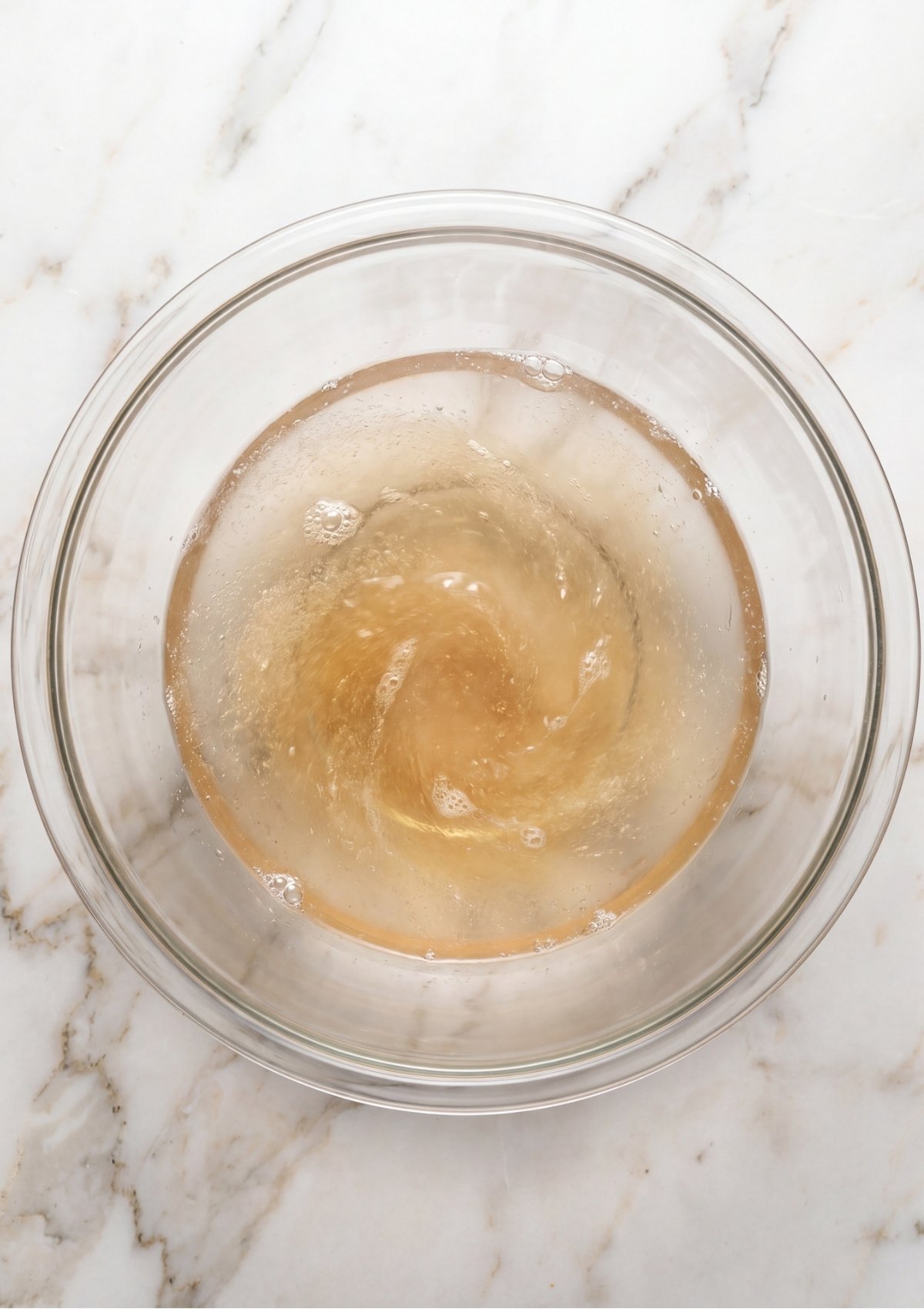 Looking down into a clear glass mixing bowl as unflavored gelatin powder is whisked into hot water, creating a slight swirl pattern.