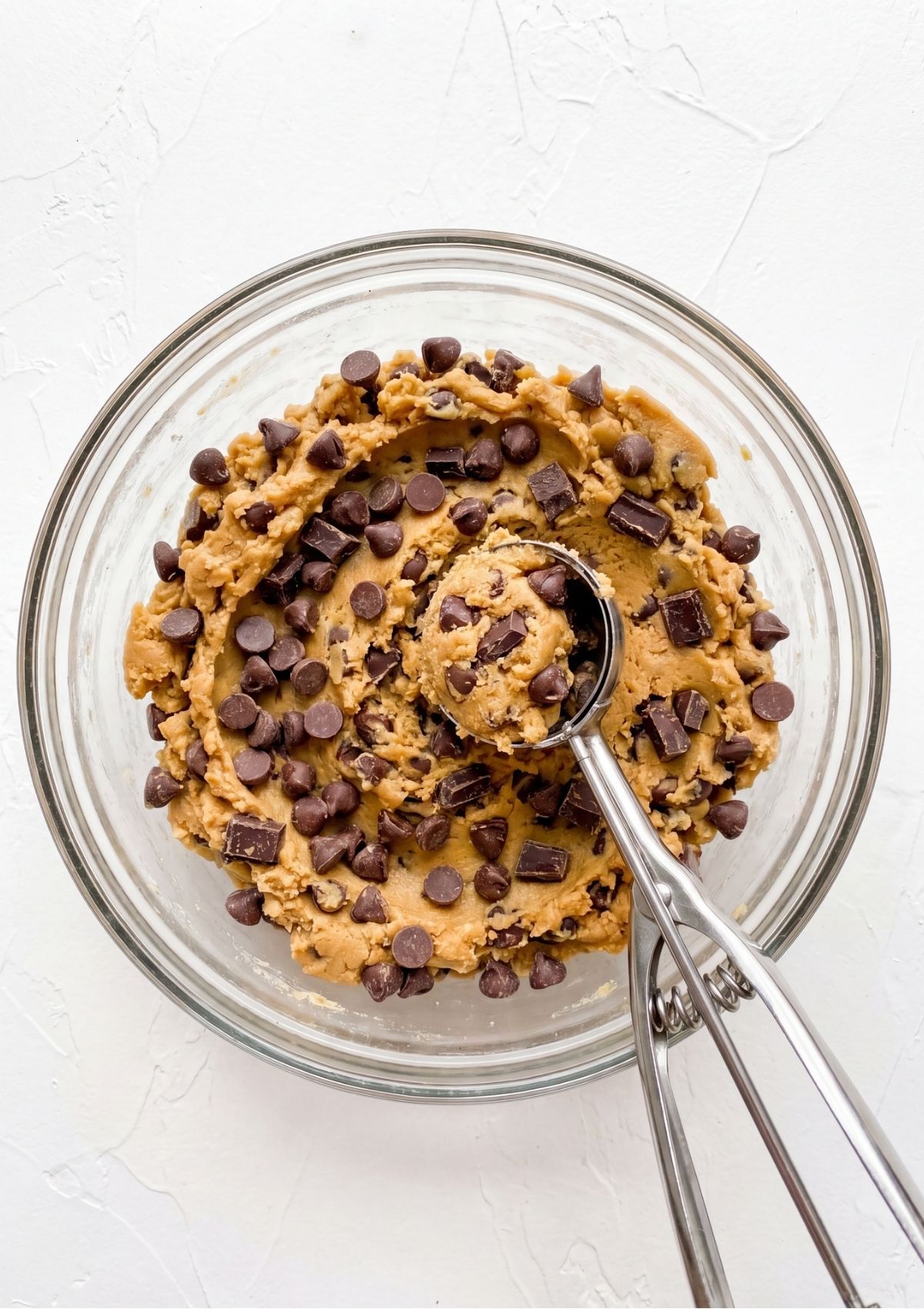 A metal cookie scoop scooping thick cookie dough loaded with chocolate chips and chunks out of a glass bowl