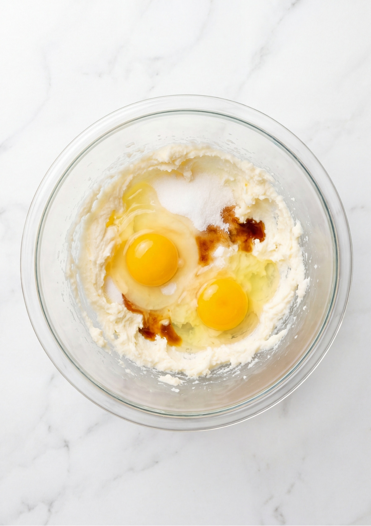Two raw eggs, white granulated sugar, and dark vanilla extract added to creamed butter in a glass mixing bowl.