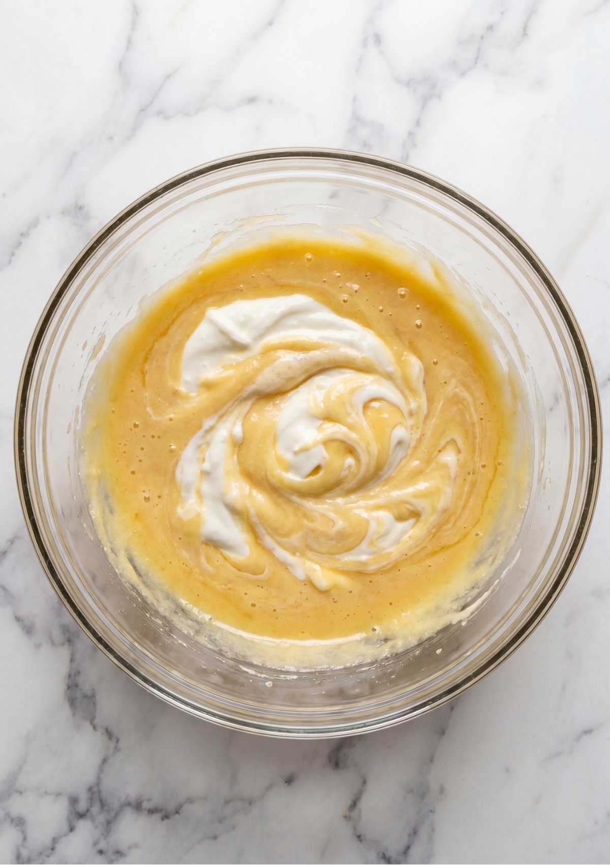 White Greek yogurt being swirled into a golden yellow cake batter in a glass mixing bowl.