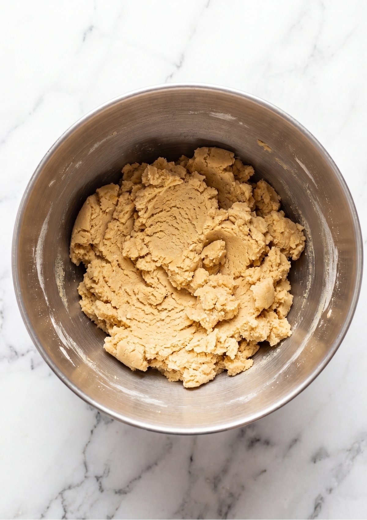 A thick, cream-colored cookie dough base without chocolate chips inside a silver mixing bowl on a marble counter