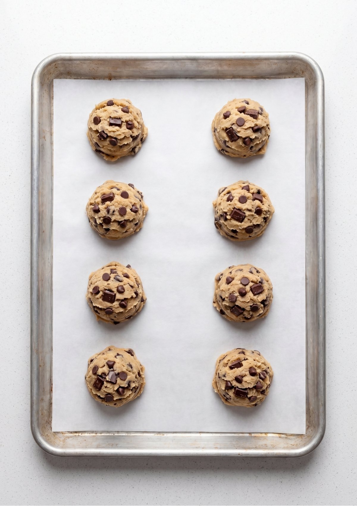 Eight large chocolate chip cookie dough balls arranged on a baking sheet lined with parchment paper