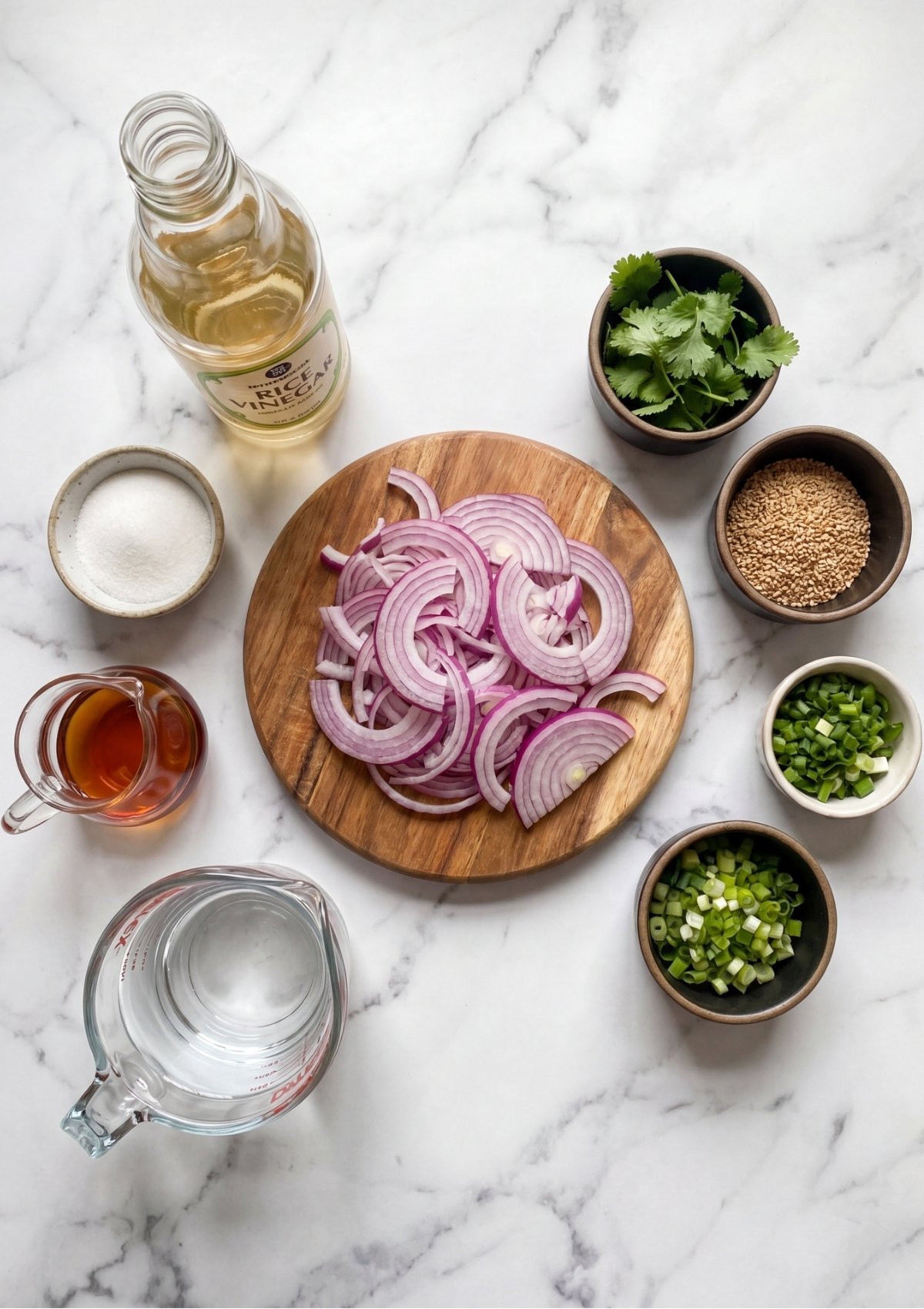An overhead view of ingredients for pickled onions arranged on a marble surface including sliced red onions on a round wooden board, rice vinegar, fish sauce, sugar, water, sesame seeds, chopped green onions, and fresh cilantro.