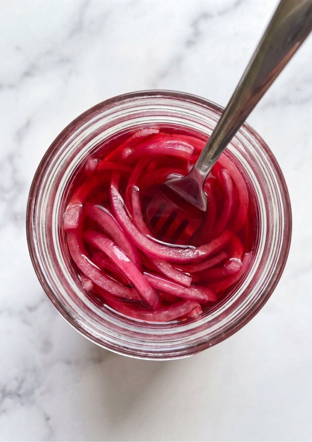 Looking down into a glass jar filled with bright pink pickled red onions in pink brine with a metal fork resting inside