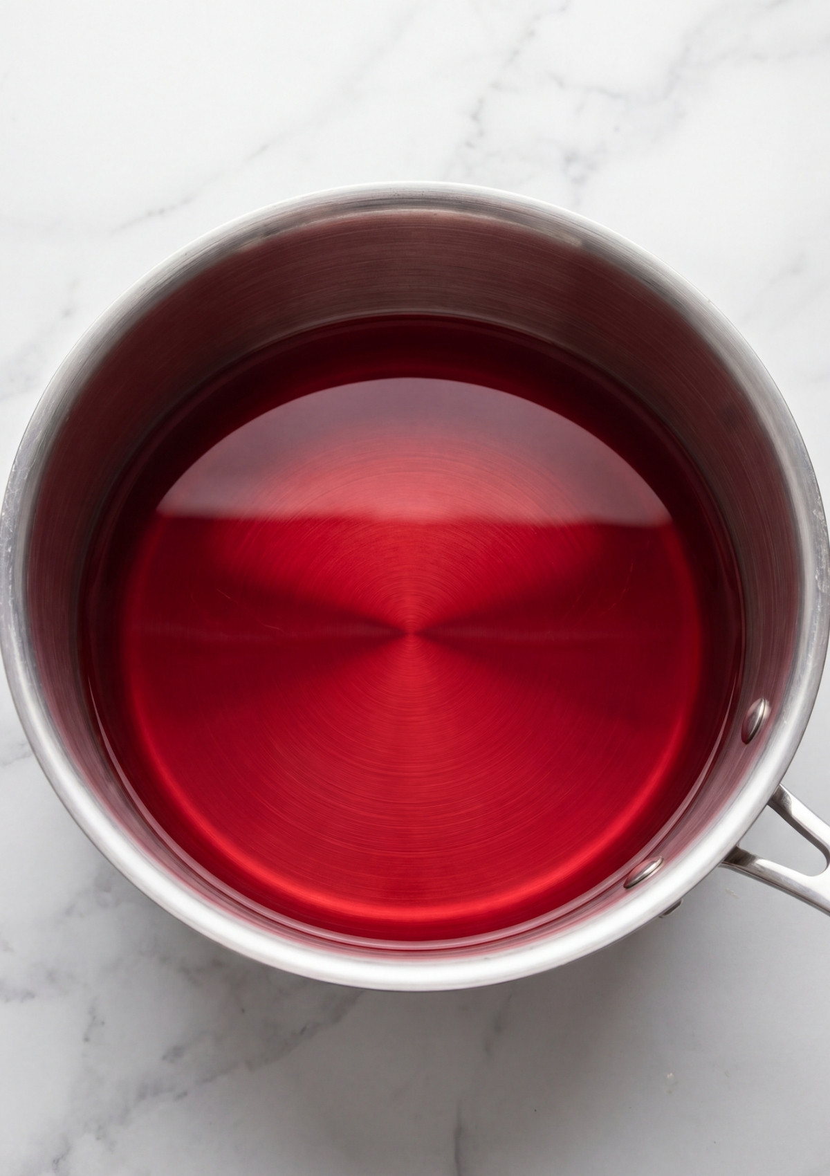A top down view of a stainless steel saucepan filled with a deep translucent red pickling liquid on a white marble background.