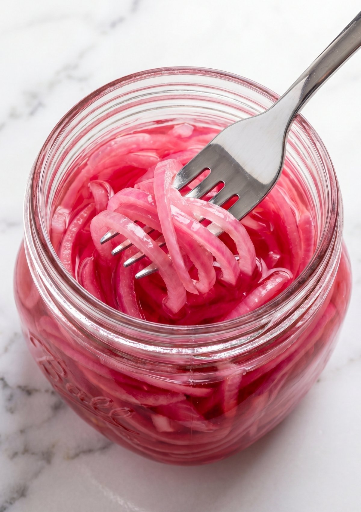 Close-up view looking down into a glass jar filled with bright pink pickled red onions being lifted out with a silver fork.