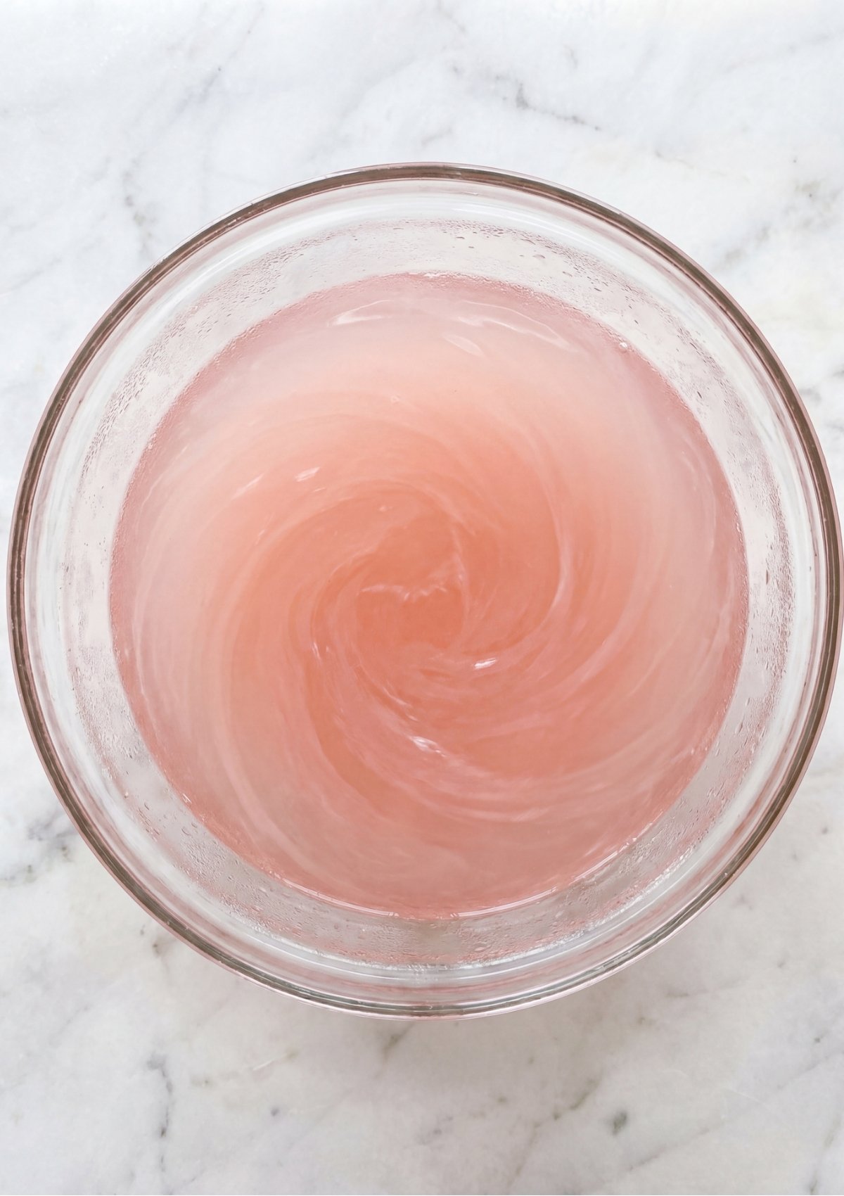 An overhead view of pink grapefruit juice being stirred into a clear glass bowl of liquid gelatin, creating a light pink swirl.