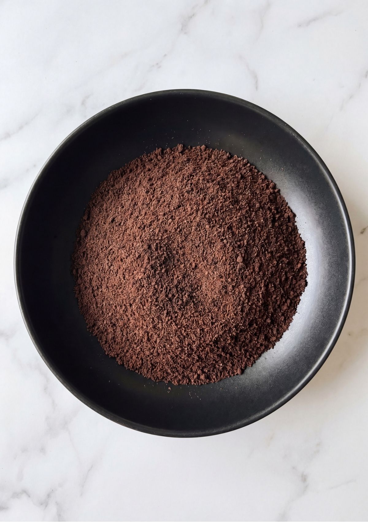 A black bowl filled with uniform, finely ground dark chocolate cookie crumbs ready for recipe assembly.
