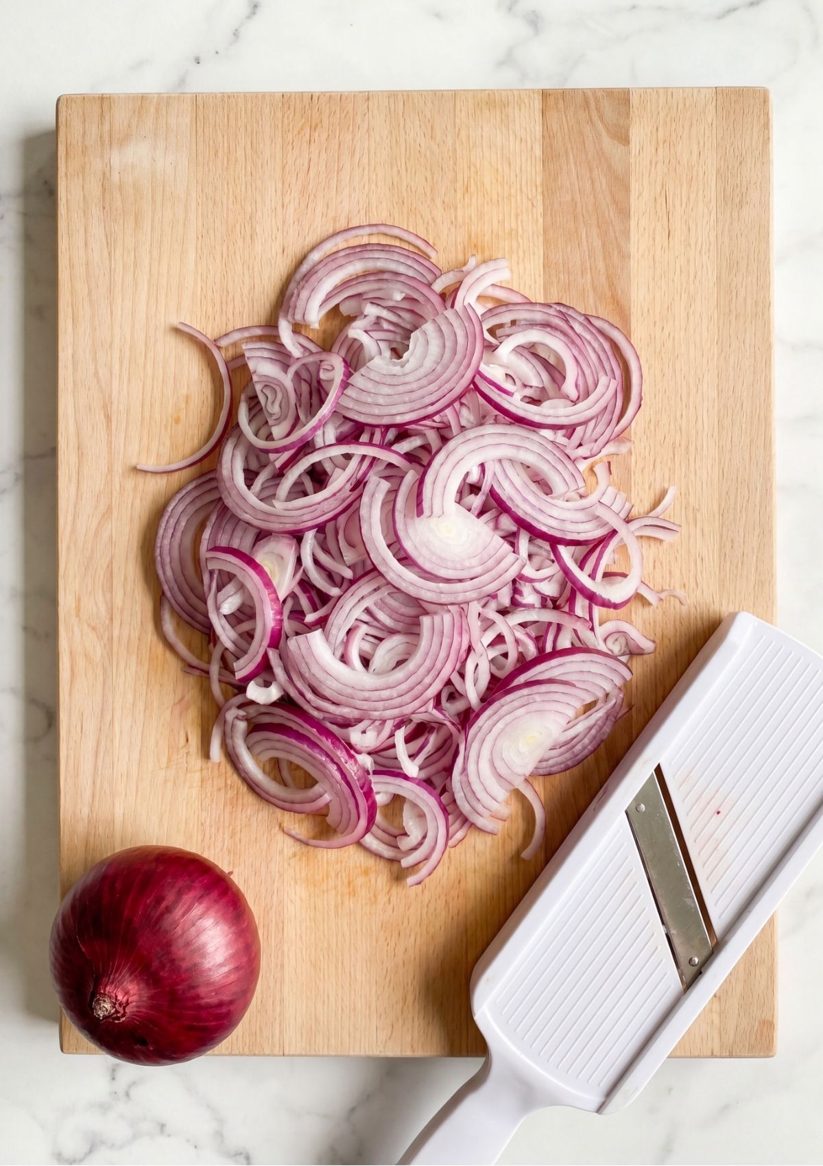 Sliced red onion rings piled on a wooden cutting board next to a whole red onion and a white mandoline slicer.