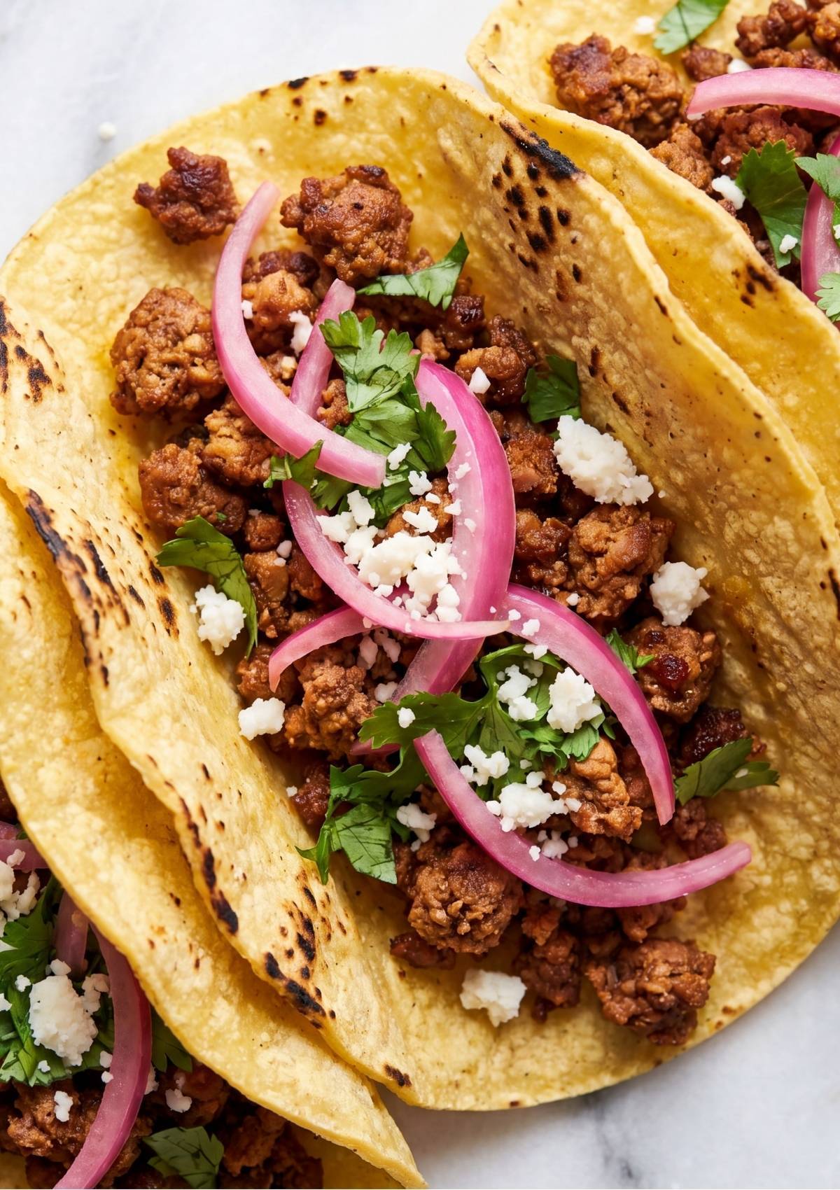 Close-up of loaded ground beef tacos in corn tortillas topped with fresh cilantro, crumbled white cheese, and pink pickled red onion strips