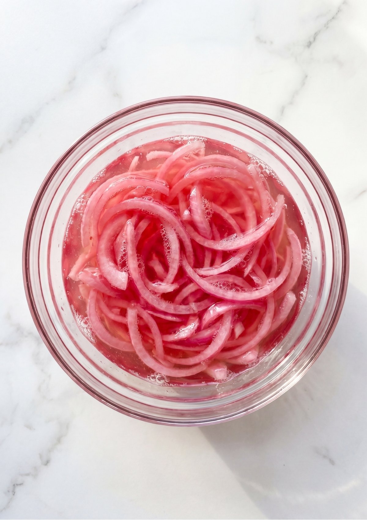 A top-down view of a clear glass bowl containing sliced red onions fully submerged in a pink vinegar pickling liquid on a white marble background.