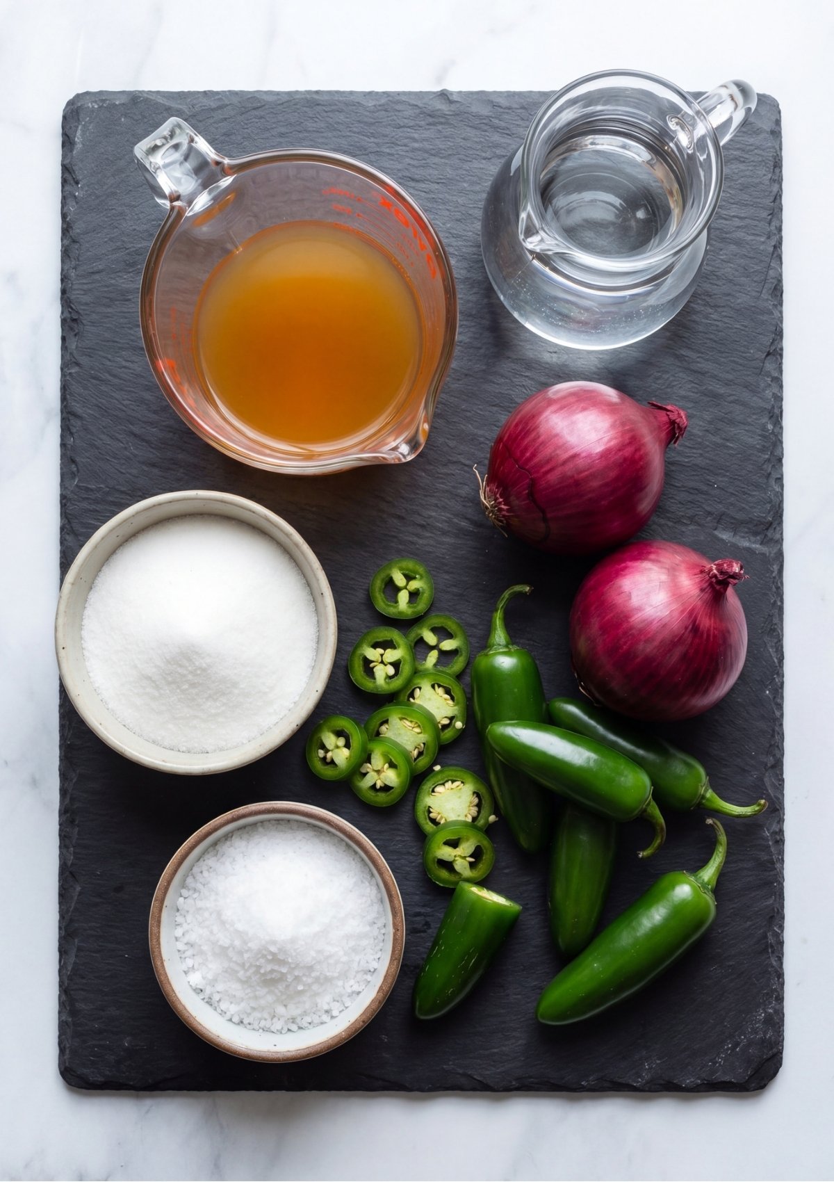 Whole red onions, fresh jalapeño peppers, sliced jalapeños, apple cider vinegar, sugar, and salt arranged on a black slate board