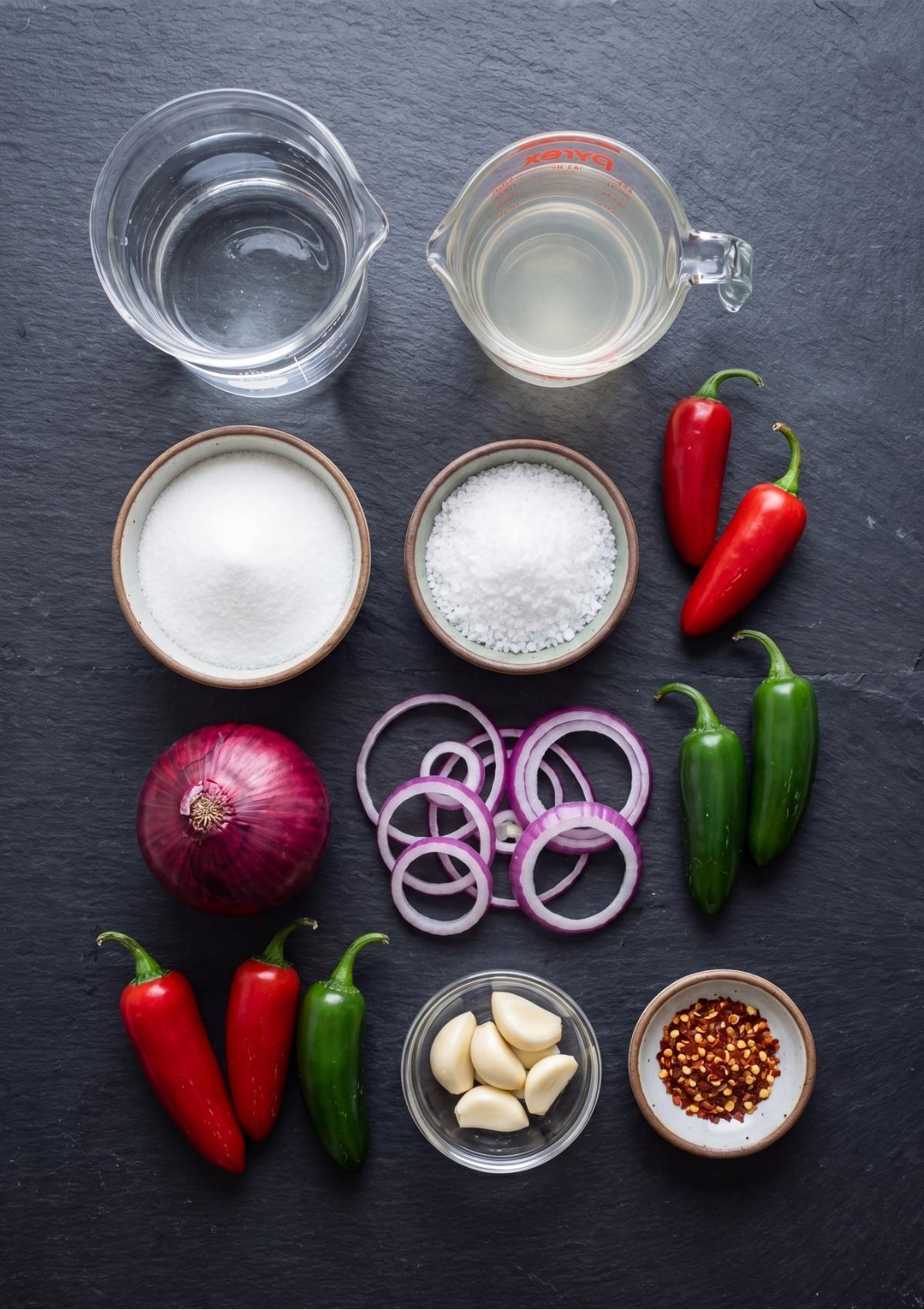 Flat lay overview of ingredients for pickled onions including a whole red onion, onion rings, vinegar, water, sugar, sea salt, garlic cloves, red pepper flakes, and fresh red and green jalapeño peppers on a dark slate surface