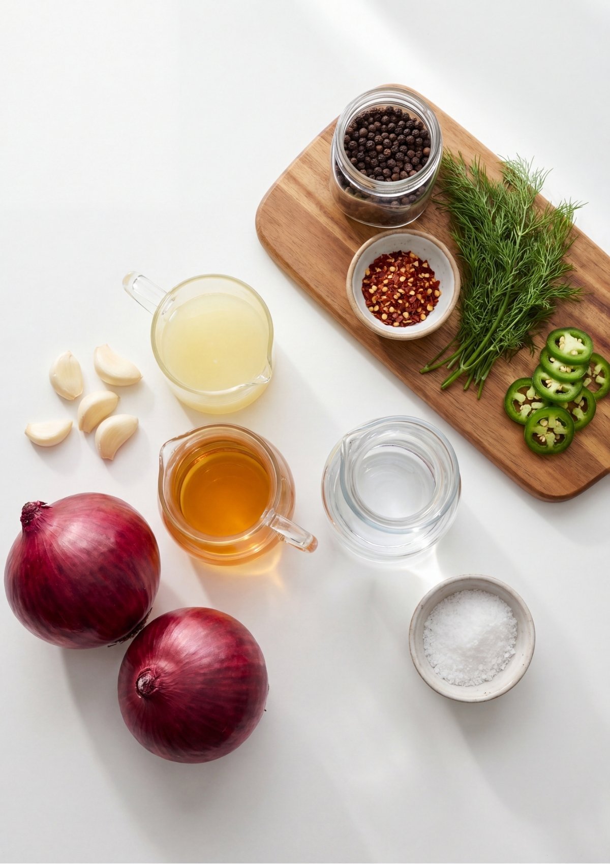 Flat lay overview of red onions, garlic cloves, vinegar, lemon juice, water, salt, peppercorns, pepper flakes, dill, and jalapeño slices on a white surface