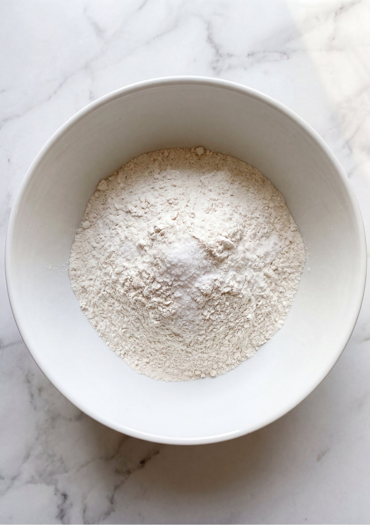 A white ceramic bowl filled with a mixture of white whole wheat and all purpose flour with a pinch of salt on top.