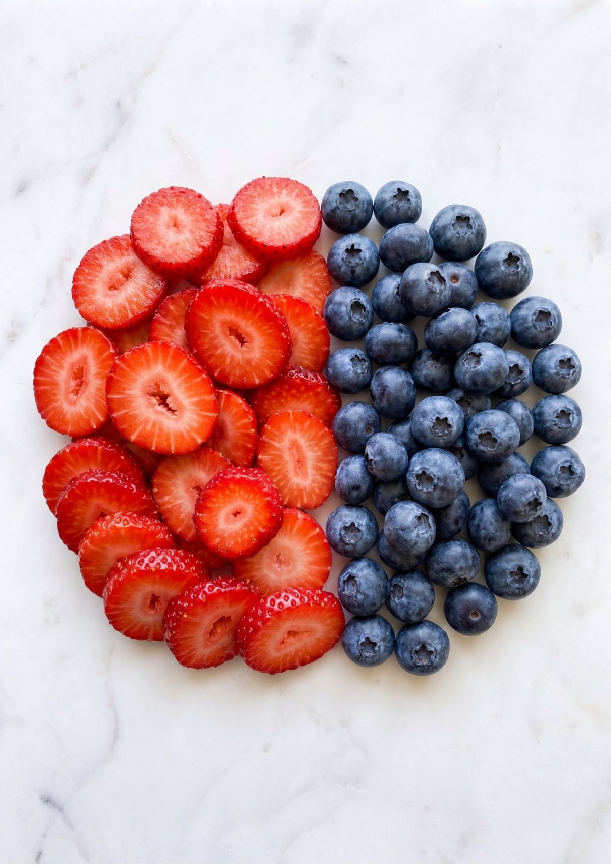 Sliced red strawberries and whole blueberries arranged in a circular shape on a white marble countertop.