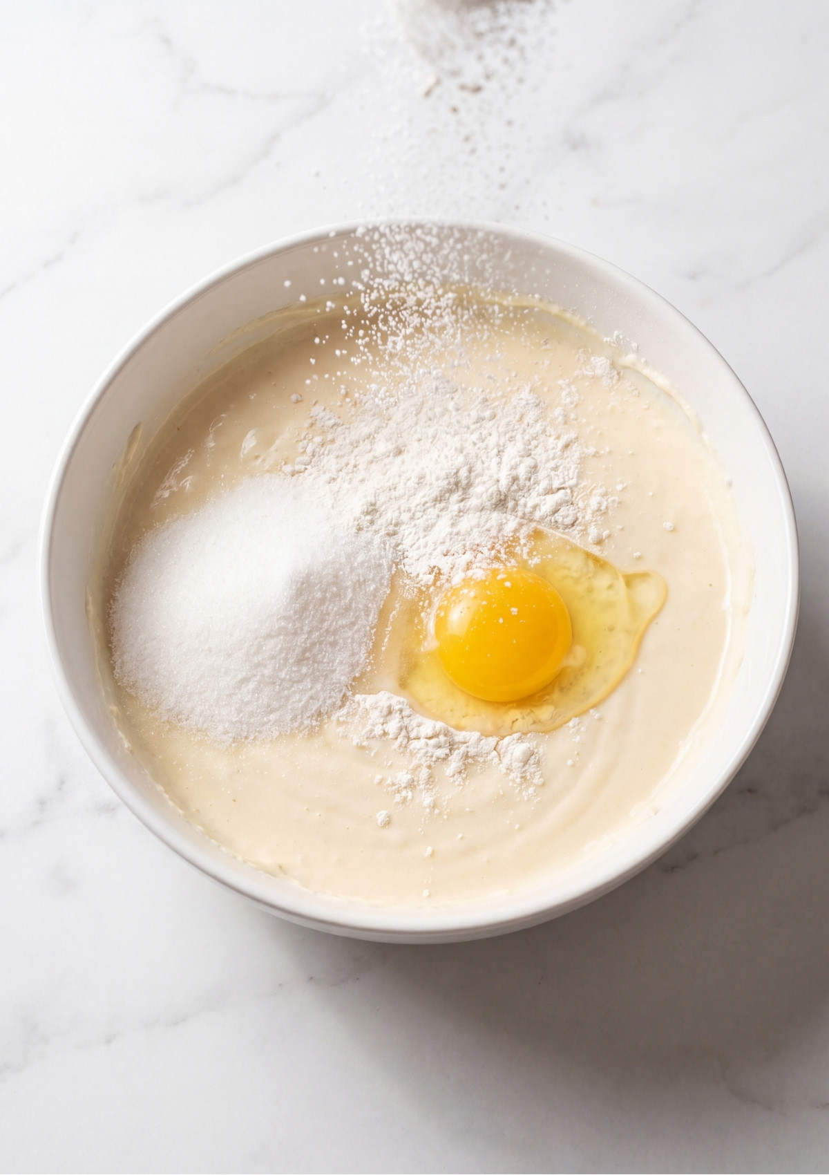 A top down view of a white bowl on a marble surface containing cheesecake batter with an egg, sugar, and flour being added.