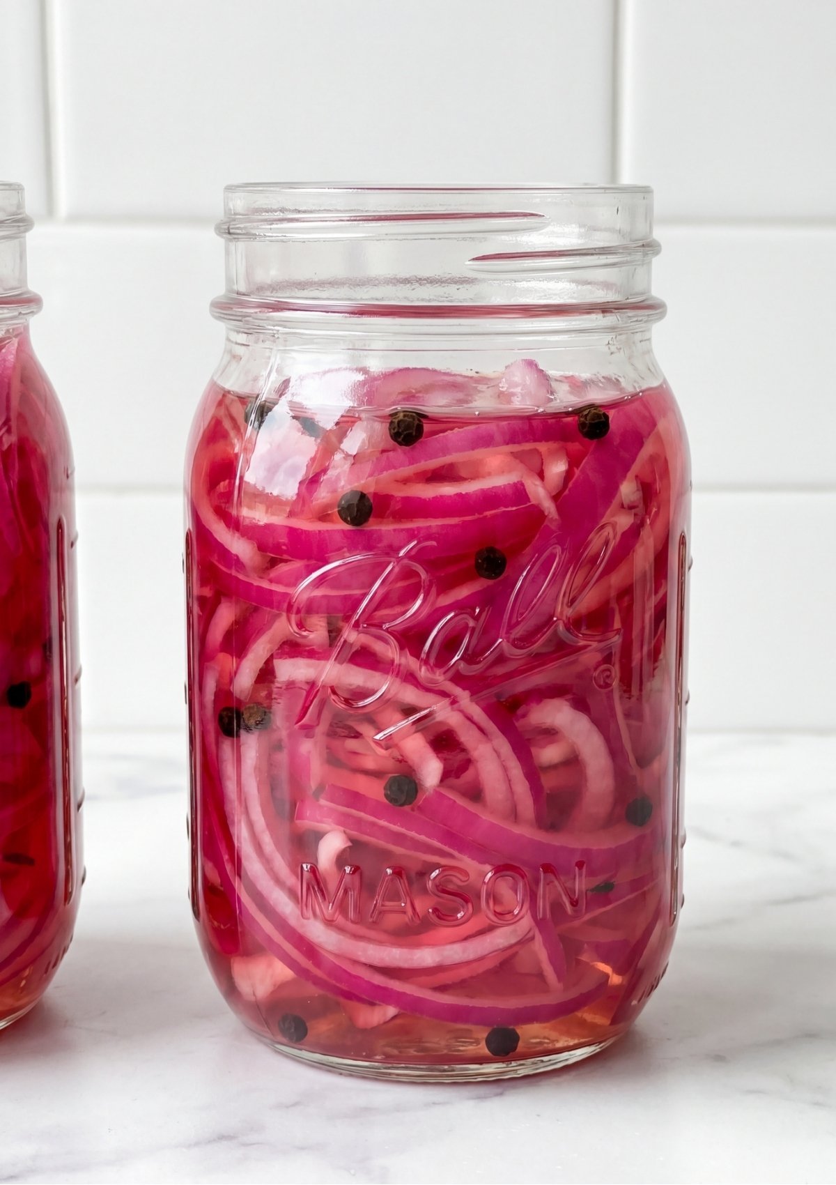 Close up view of a glass mason jar filled with bright pink pickled red onion slices and black peppercorns in clear brine.
