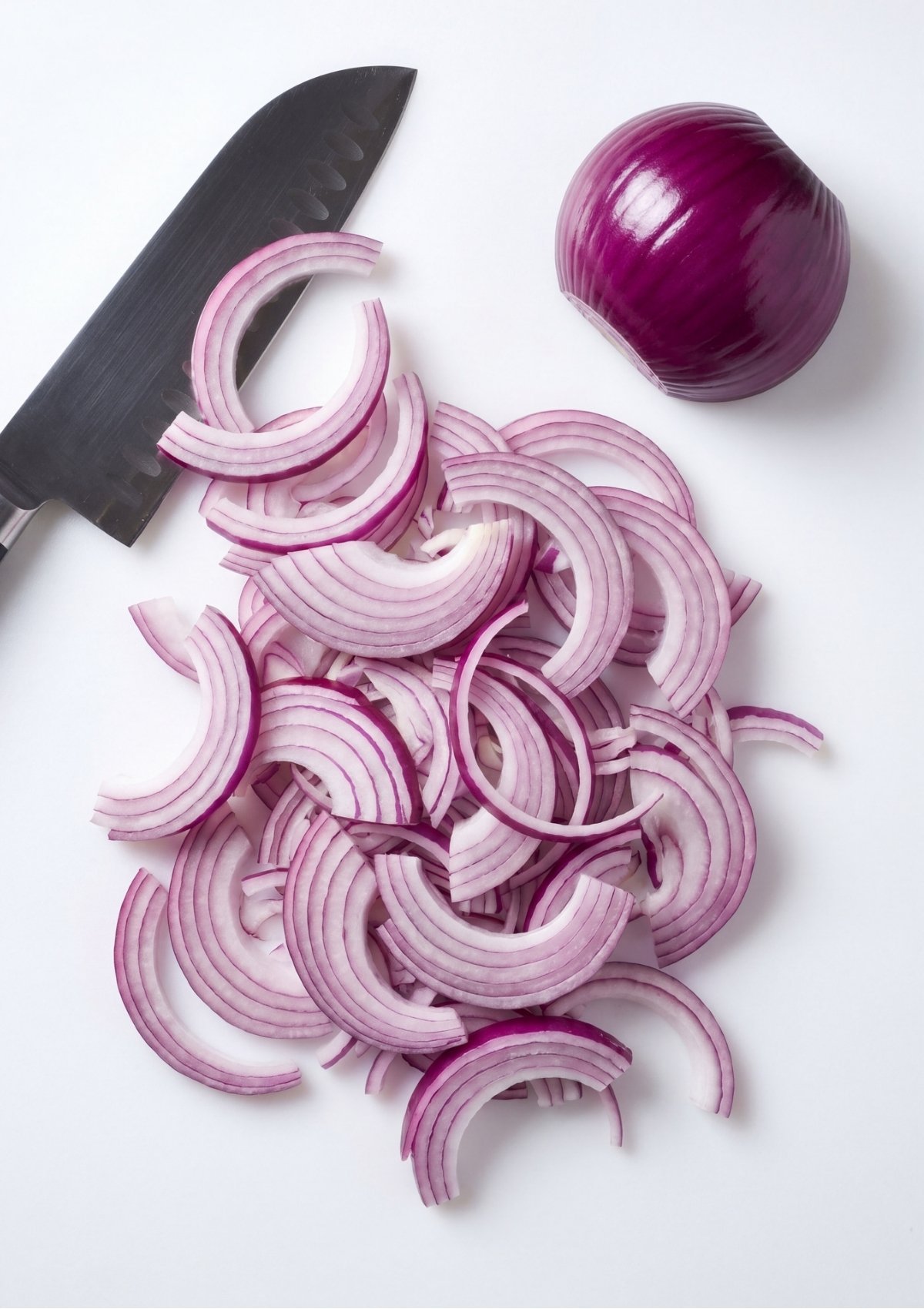 Thinly sliced half-moons of red onion piled on a white cutting board next to a chef knife and half of a peeled red onion