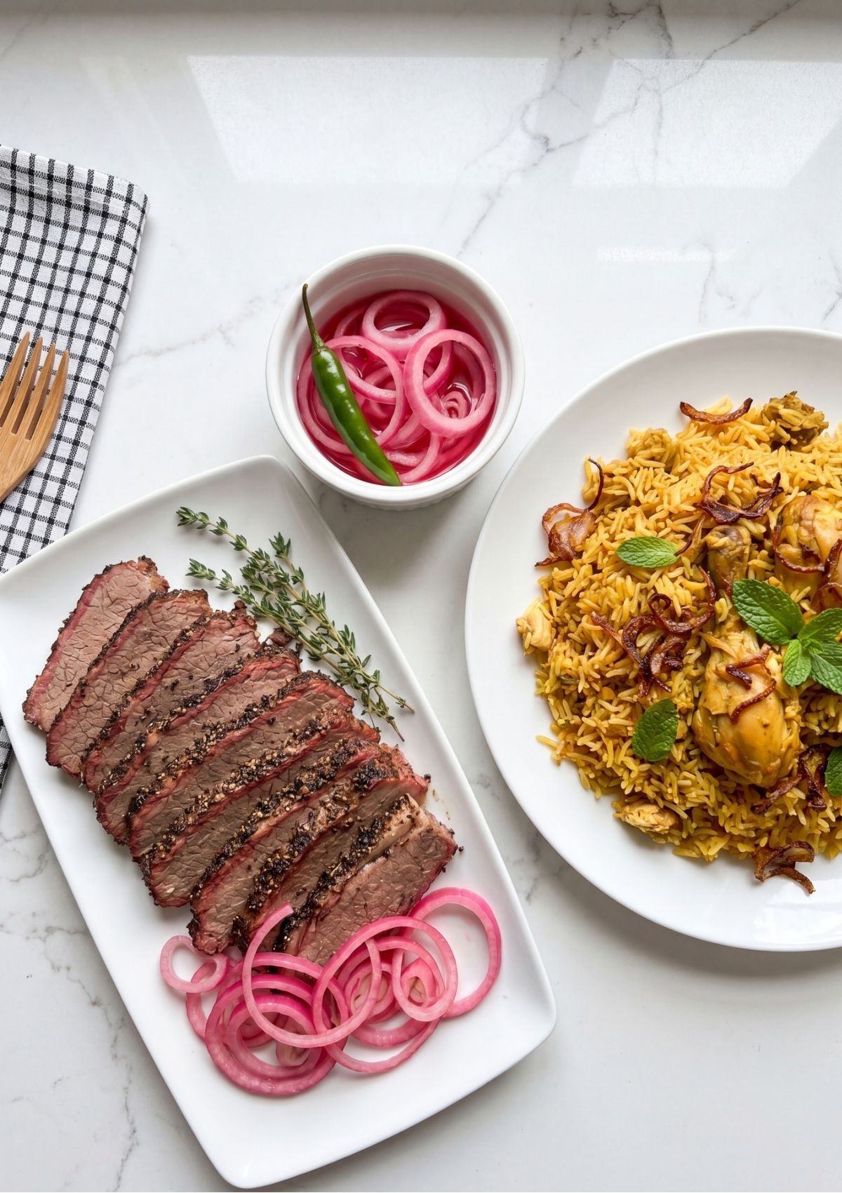 An overhead view of a meal spread featuring a platter of sliced beef brisket, a plate of chicken biryani rice, and a small bowl of pink pickled onions with a green chili