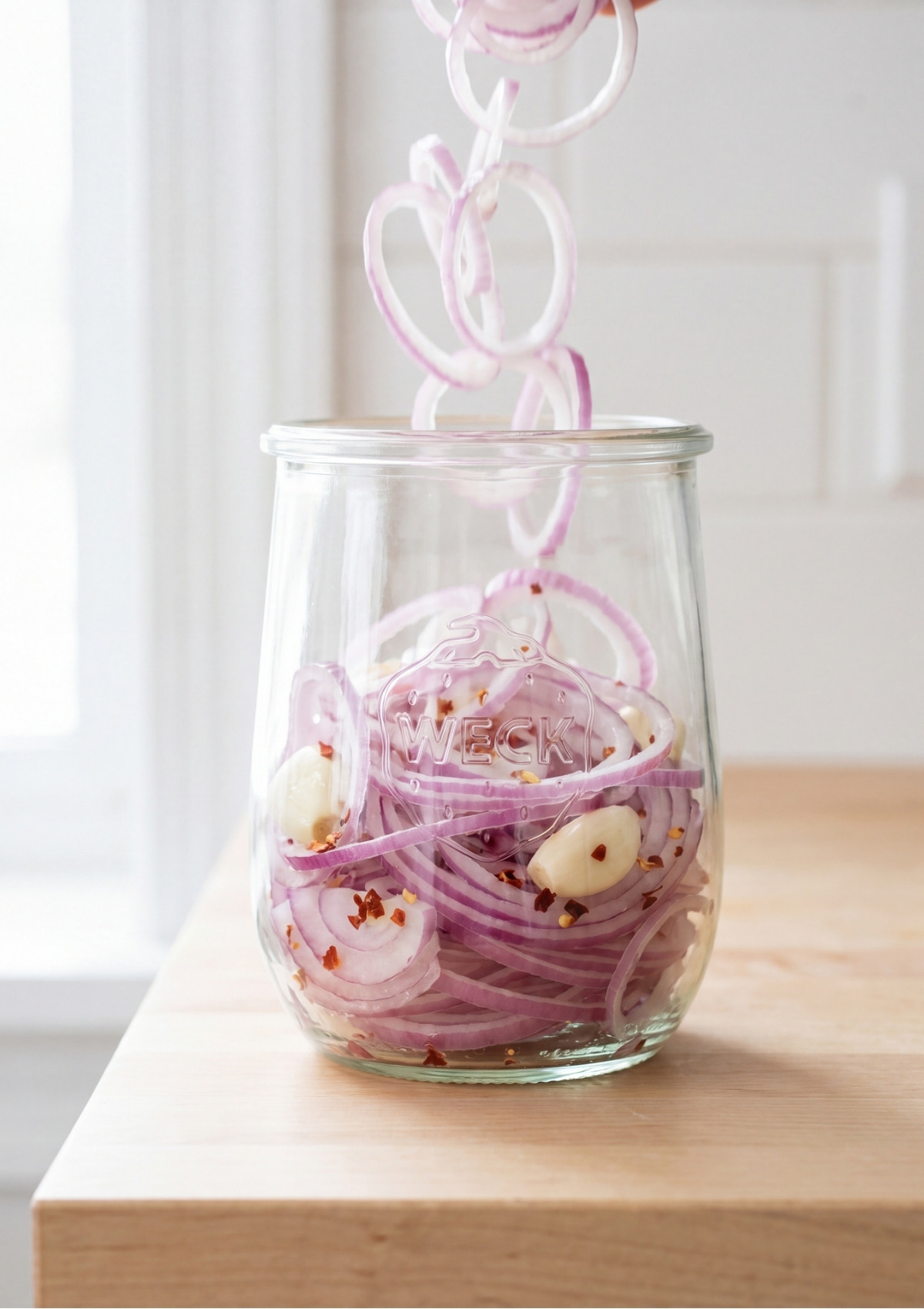 Thinly sliced red onions being dropped into a clear glass jar containing fresh garlic cloves and red pepper flakes
