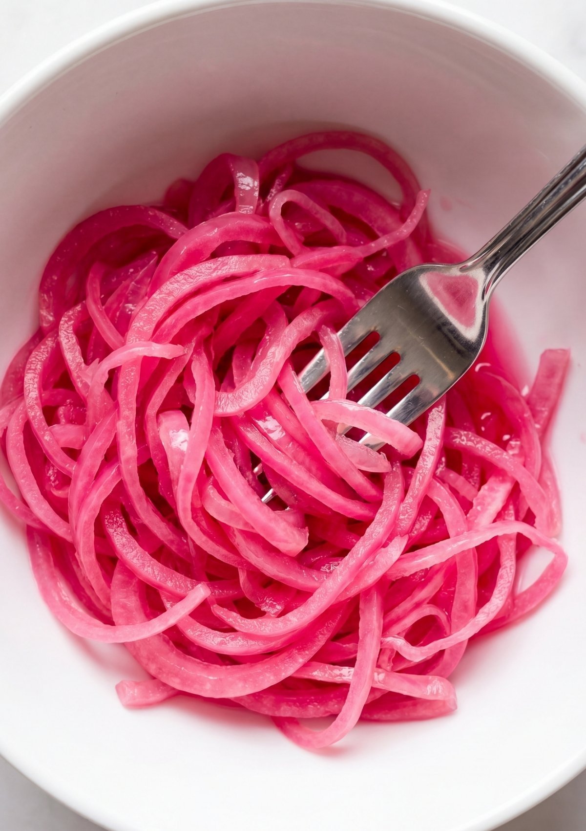 A high angle view of a white ceramic bowl filled with bright pink pickled red onion slices being twirled on a silver fork.