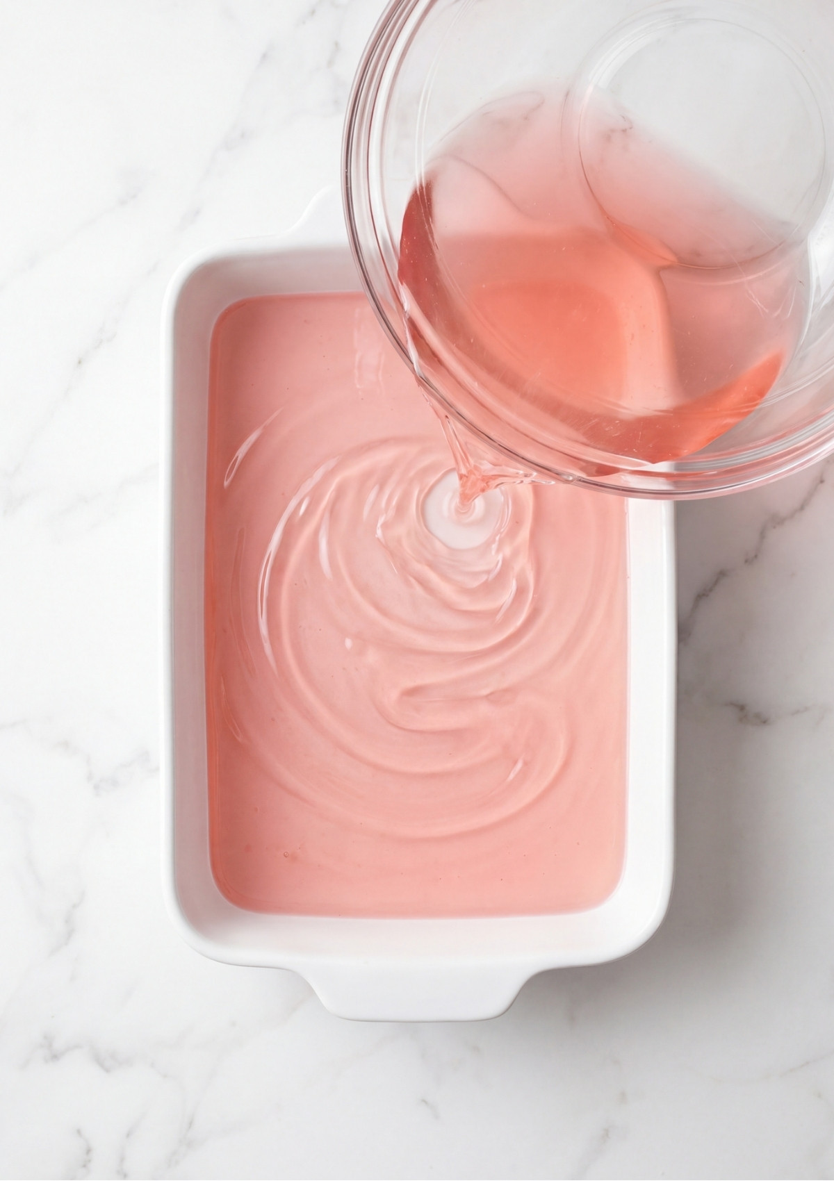 Liquid pink gelatin being poured from a glass bowl into a white ceramic rectangular baking dish on a white marble countertop.
