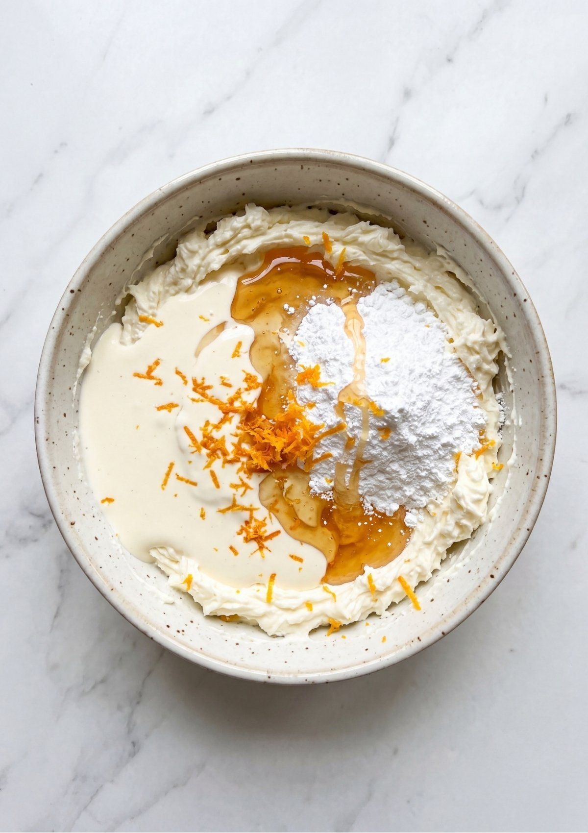 A bowl containing whipped cream topped with honey, powdered sugar, and lemon zest before being folded together for a Patriotic Tartlets Recipe for Summer Parties.