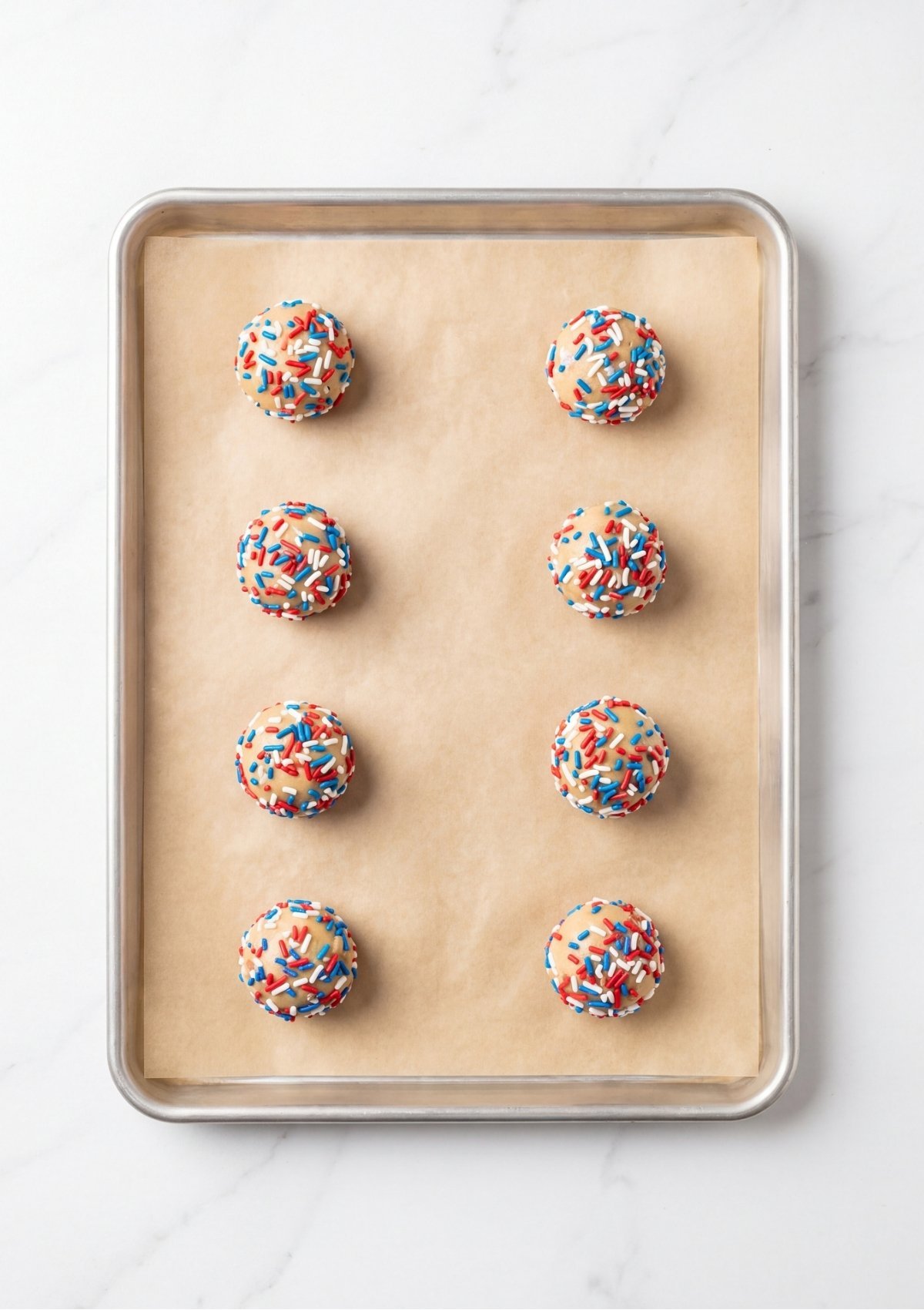 Eight balls of cookie dough coated in red white and blue sprinkles on a parchment lined baking sheet