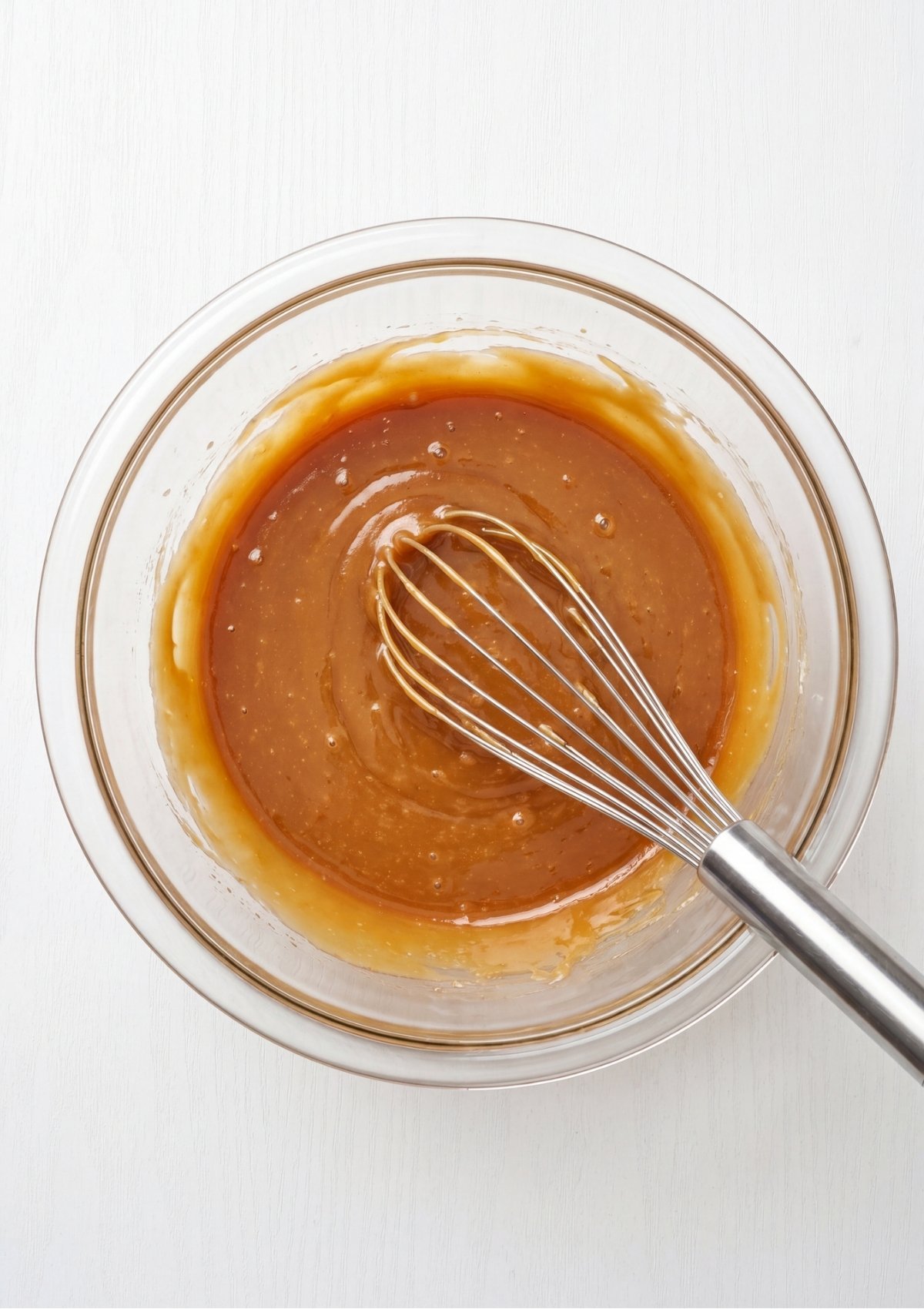 A metal whisk resting inside a glossy caramel colored mixture of melted butter and dissolved sugars within a clear glass bowl