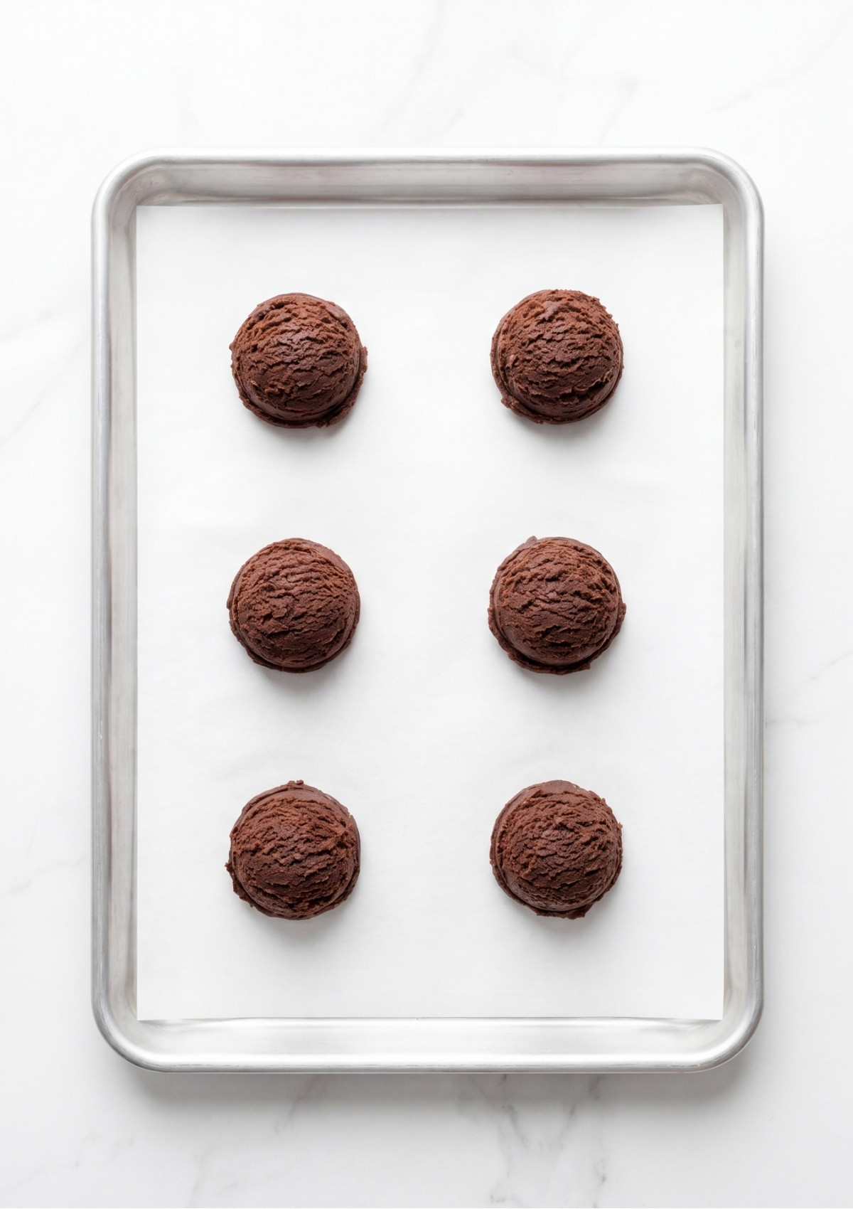 Six perfectly rounded scoops of chocolate cookie dough arranged on a parchment paper lined aluminum baking sheet.