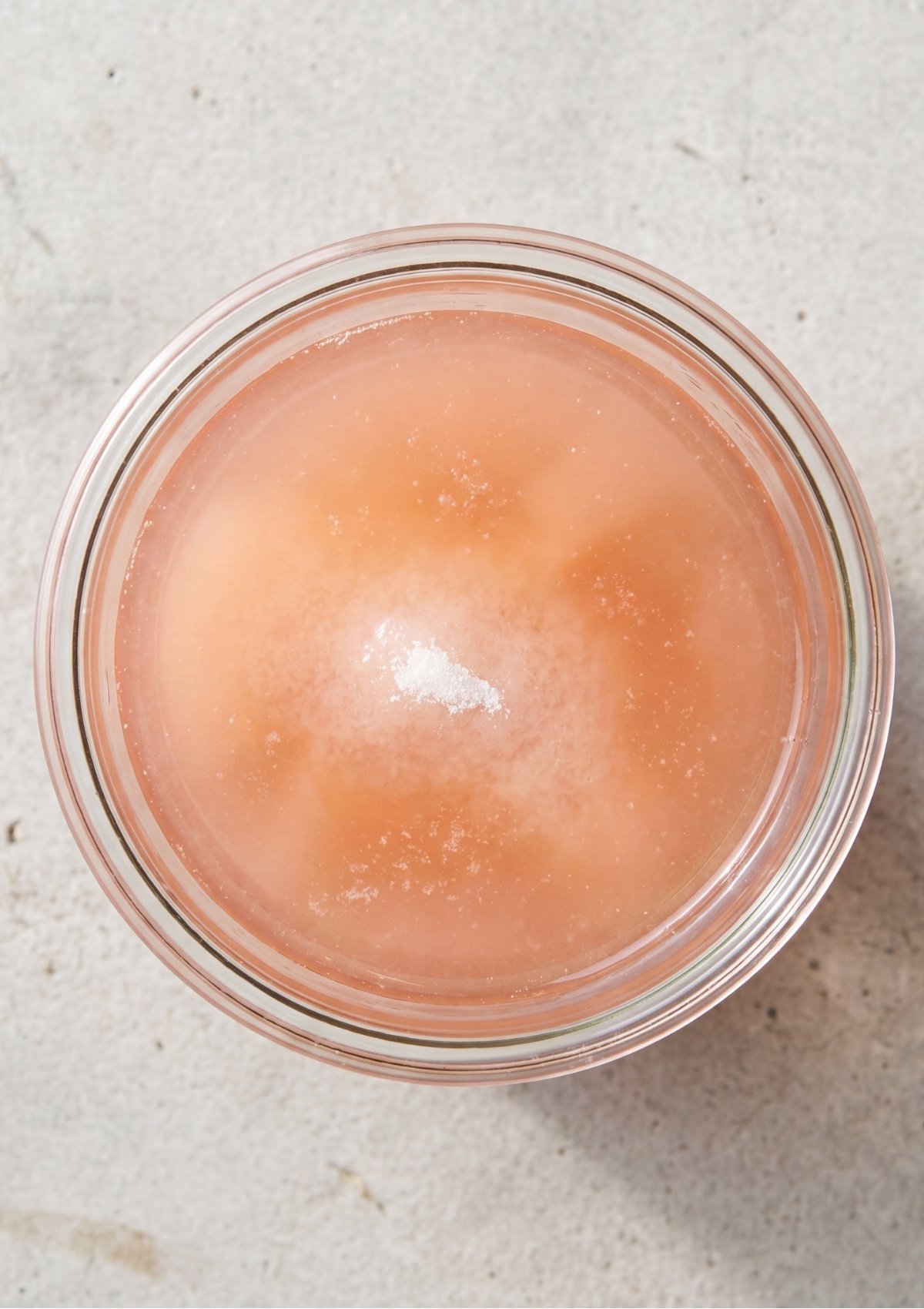 Top-down view of a clear glass jar filled with a light pink translucent pickling liquid with grains of salt and sugar settling at the bottom