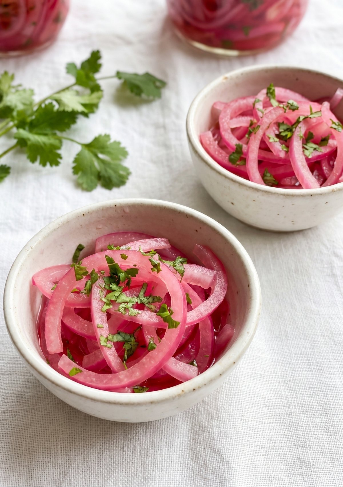 Two small white ceramic bowls filled with bright pink pickled red onions garnished with fresh chopped cilantro, set on a white textured tablecloth next to extra cilantro sprigs.
