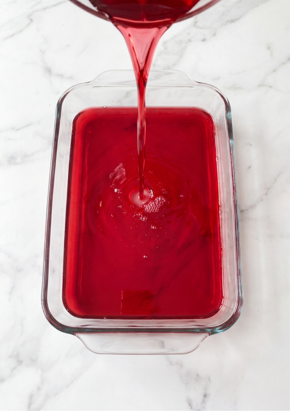 Liquid red gelatin mixture being poured from a pot into a clear rectangular glass baking dish