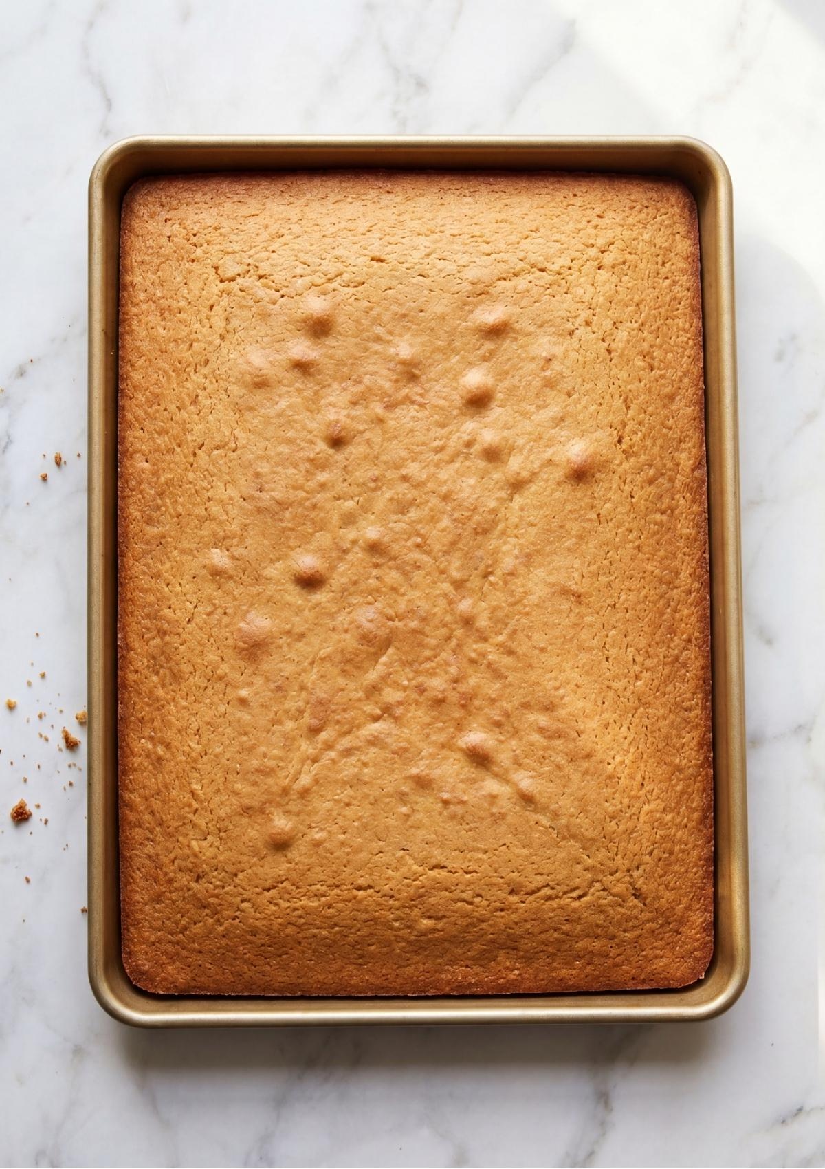 A golden brown rectangular sheet cake baked in a metallic baking pan resting on a marble countertop.