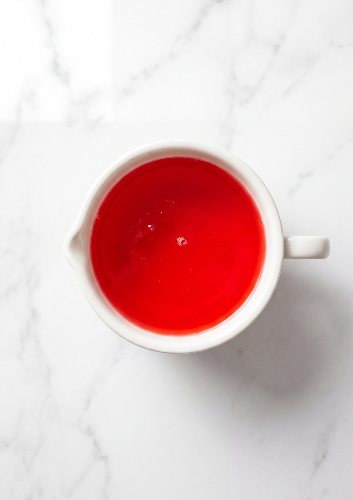 A top down view of bright red strawberry liquid gelatin in a white ceramic measuring cup on a marble surface
