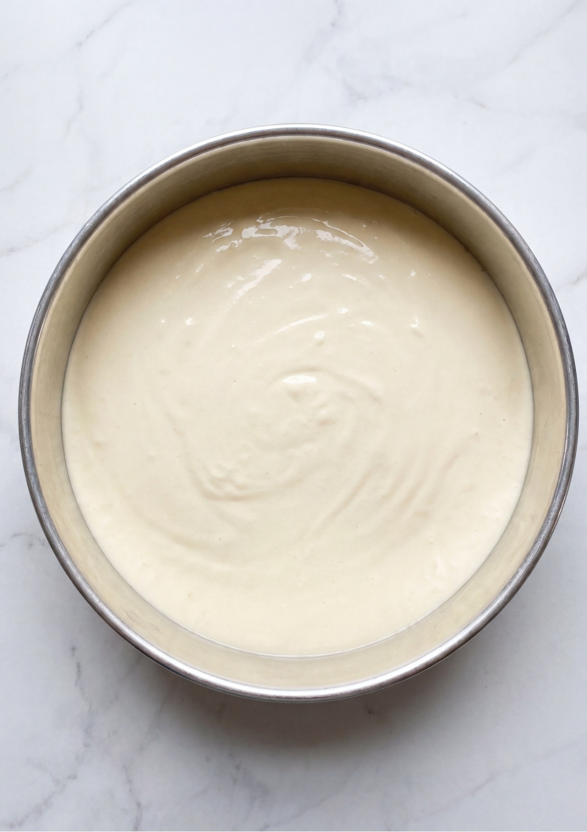 A silver round cake pan filled with smooth cheesecake batter ready to be baked on a marble surface.