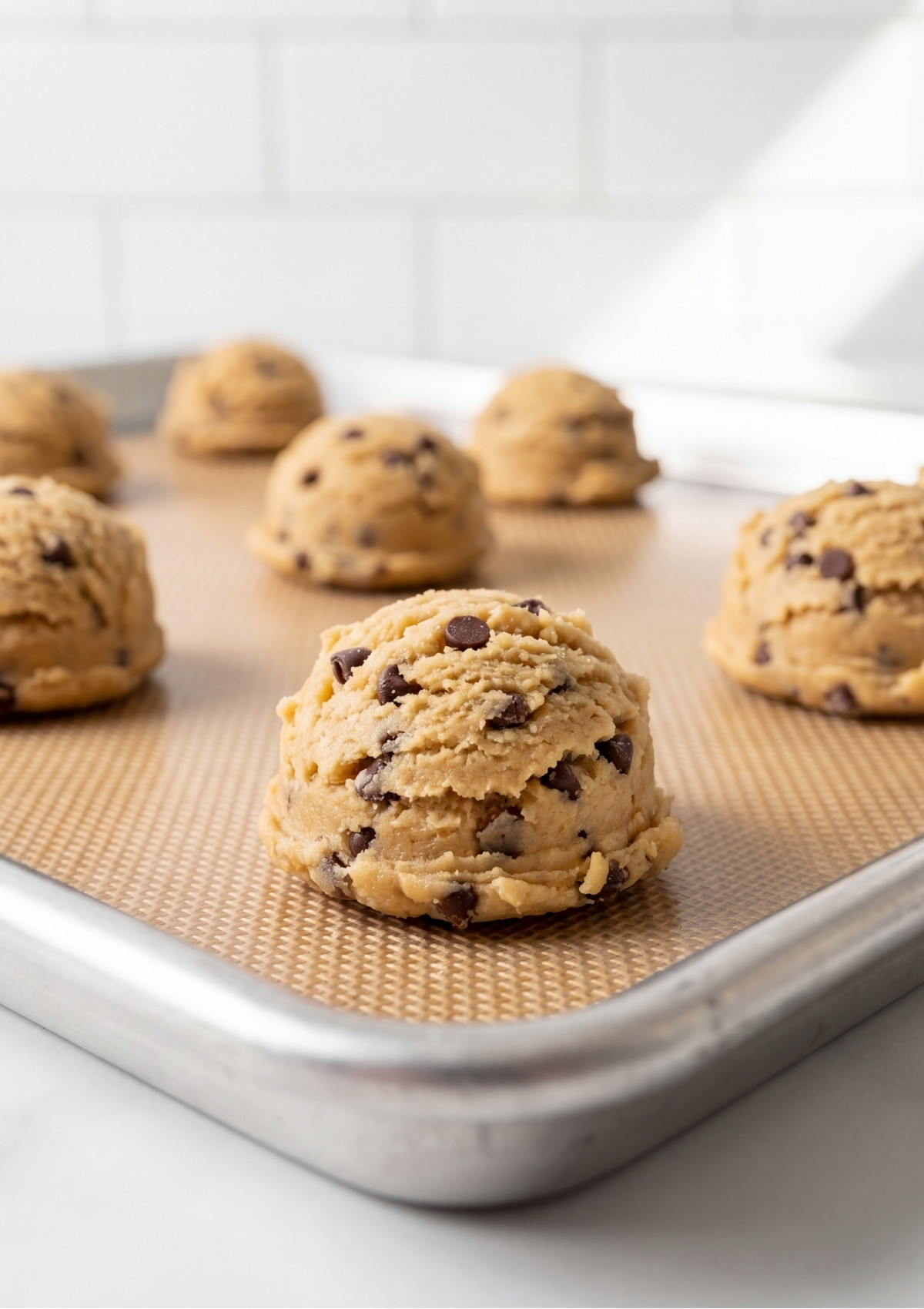 Large scoops of chocolate chip cookie dough arranged on a silicone baking mat lined sheet pan