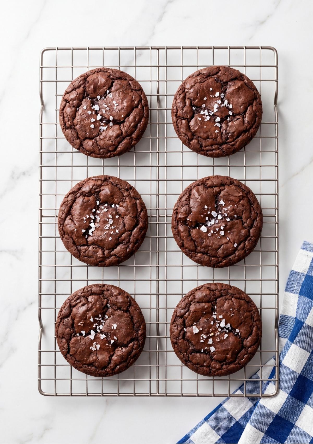 Overhead view of six baked chocolate cookies topped with flaky sea salt resting on a rectangular wire cooling rack.