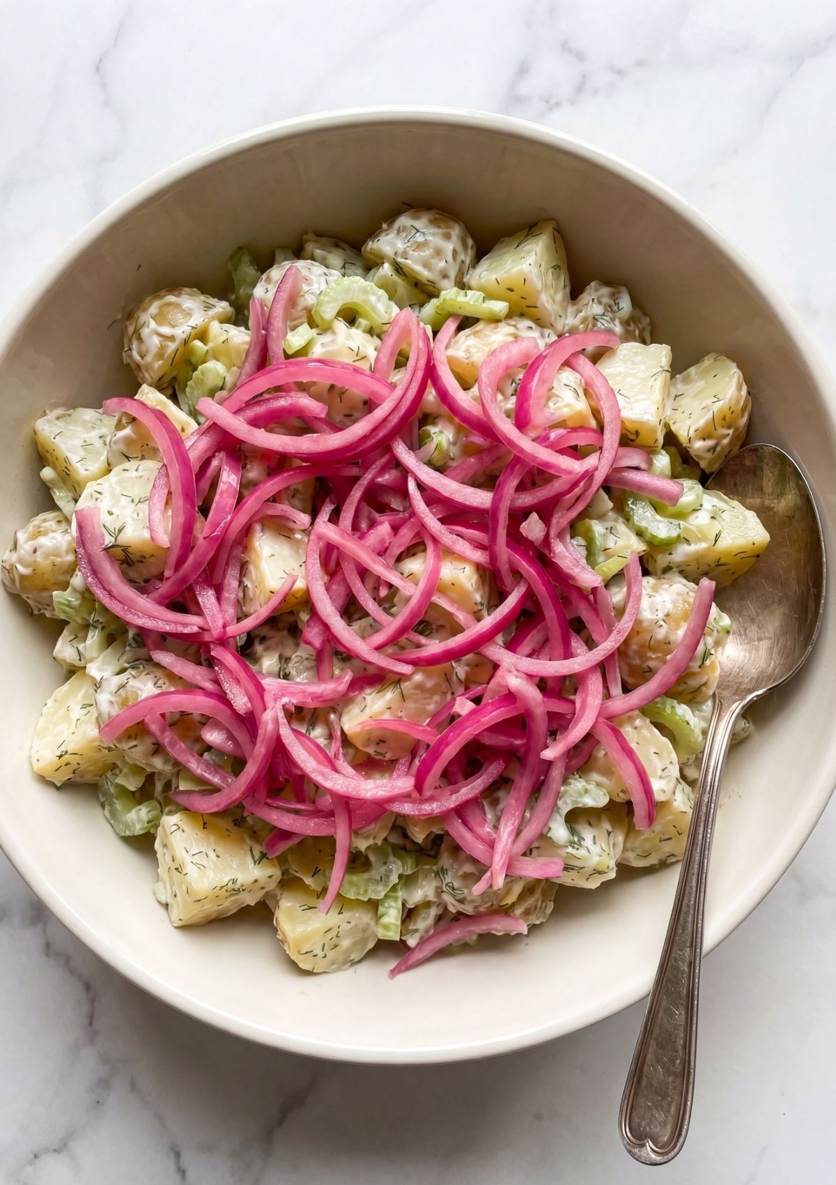 A large white serving bowl filled with creamy potato salad with celery and fresh dill topped with bright pink pickled red onions
