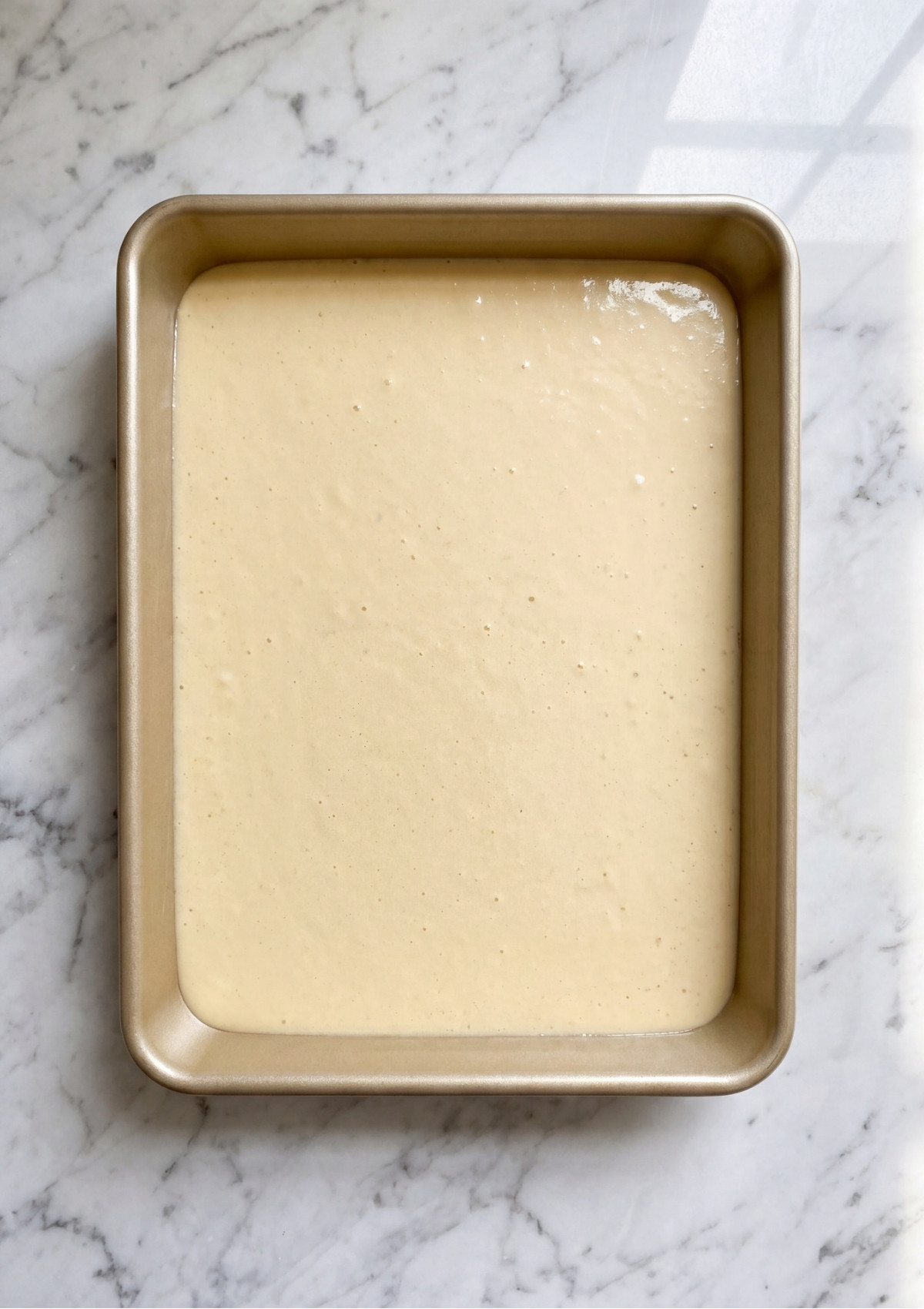 Raw vanilla cake batter spread evenly into a gold rectangular sheet pan before baking.