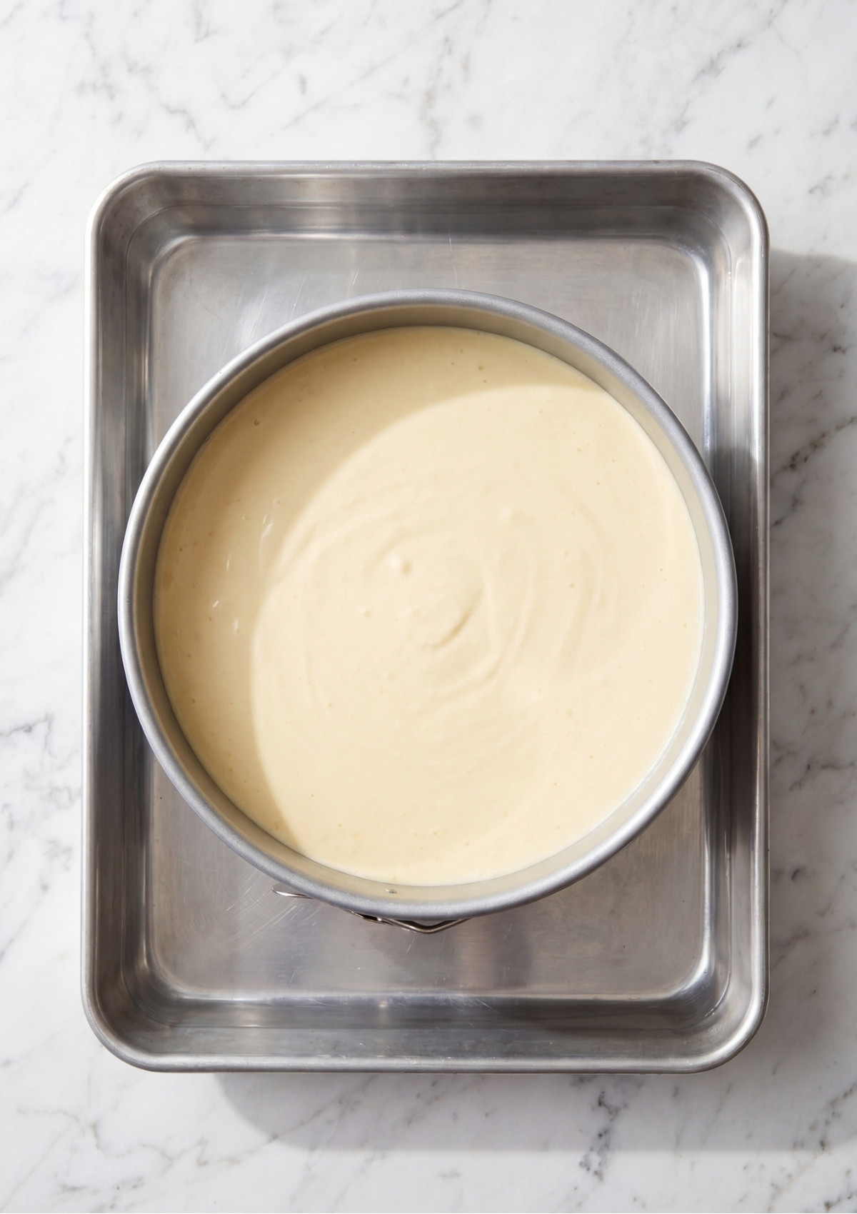 A round metal cake pan filled with pale cheesecake batter sitting inside a larger rectangular metal baking pan on a marble countertop.