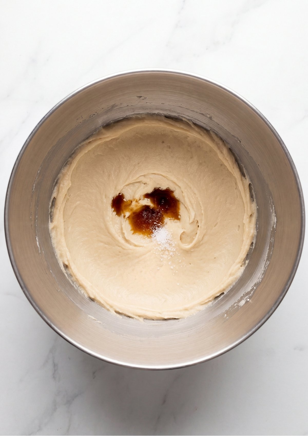 Overhead view of vanilla extract and sea salt added to a smooth butter and sugar mixture in a metal mixing bowl
