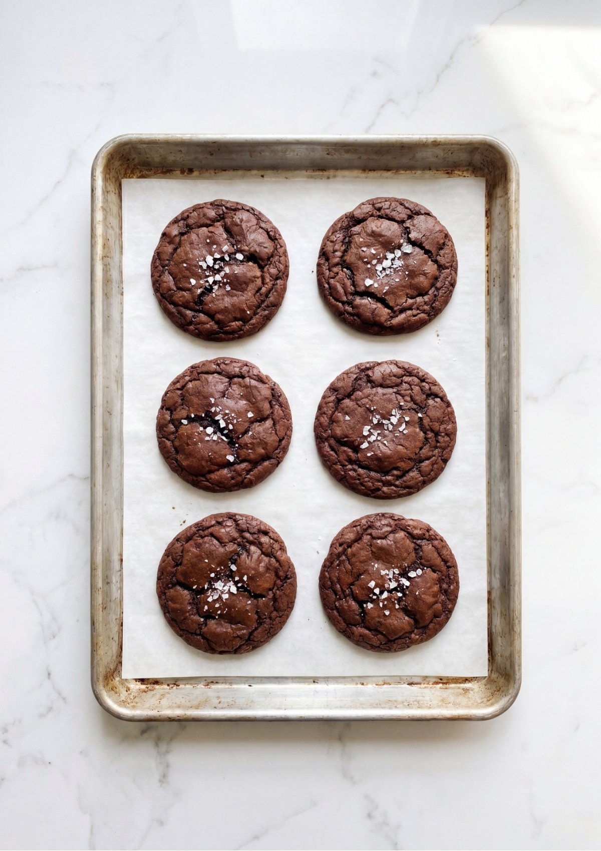 Top-down view of six freshly baked chocolate cookies with crinkly tops and a sprinkle of sea salt on a parchment-lined baking sheet.